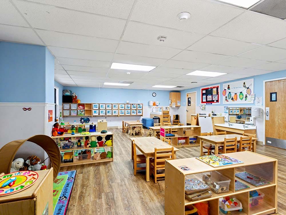 A classroom with small tables and chairs, shelves with toys, and artwork on the walls