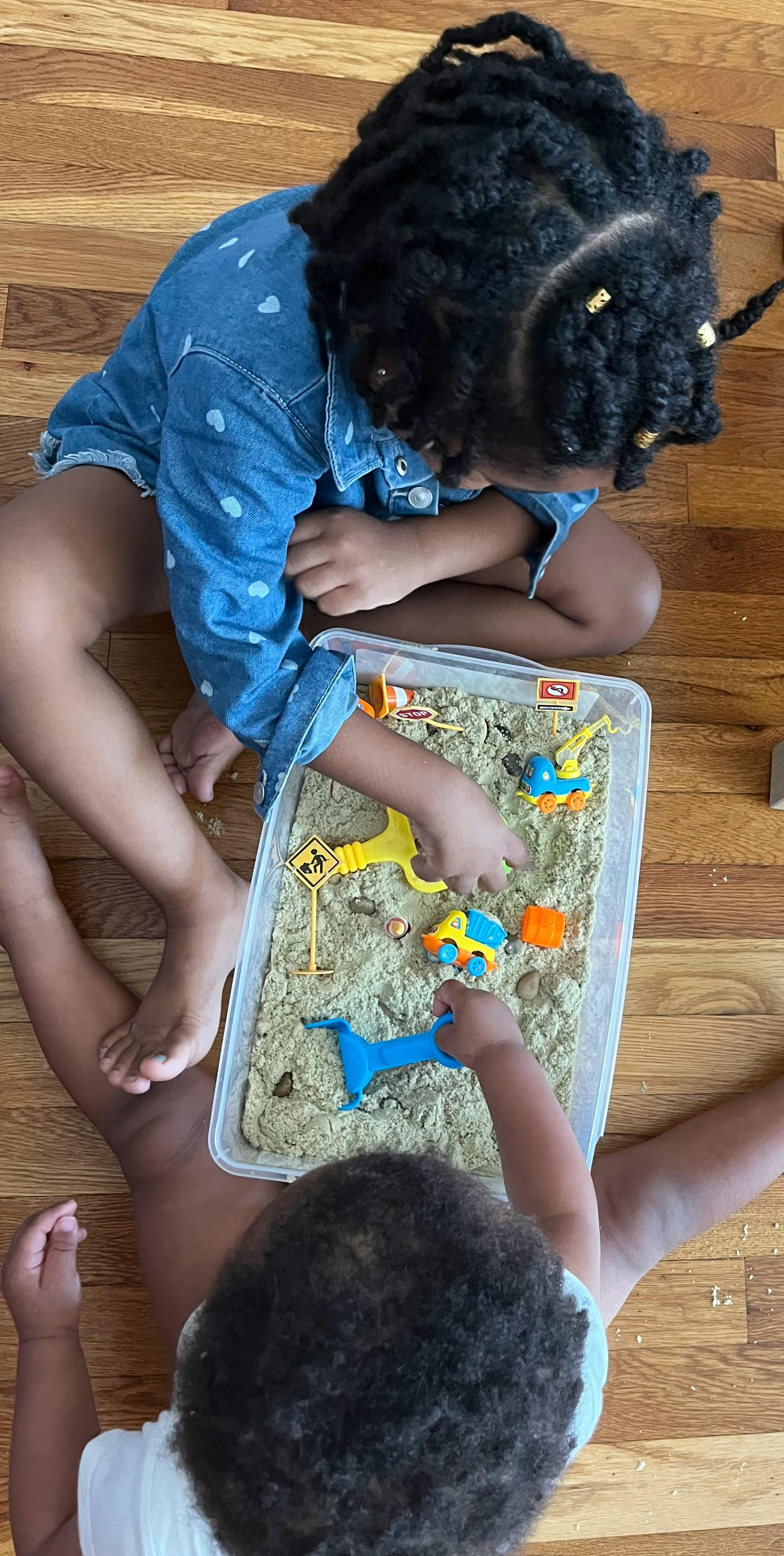 Two children playing with sand and toy construction vehicles in a clear bin