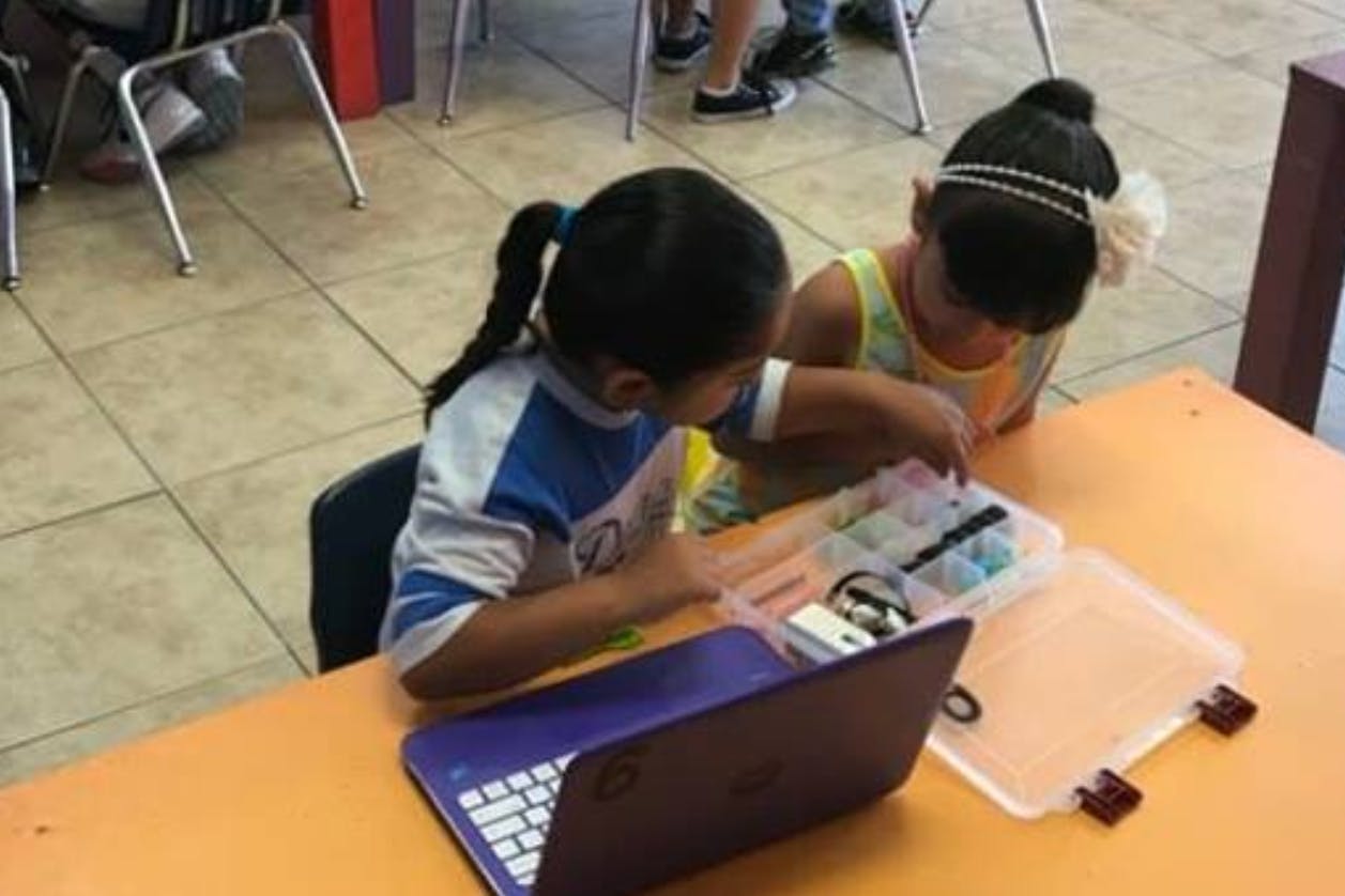 Two children looking at items in a clear container on a desk with a laptop