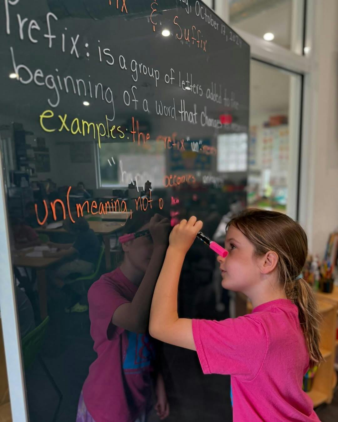 Child writing on a clear board with a pink marker