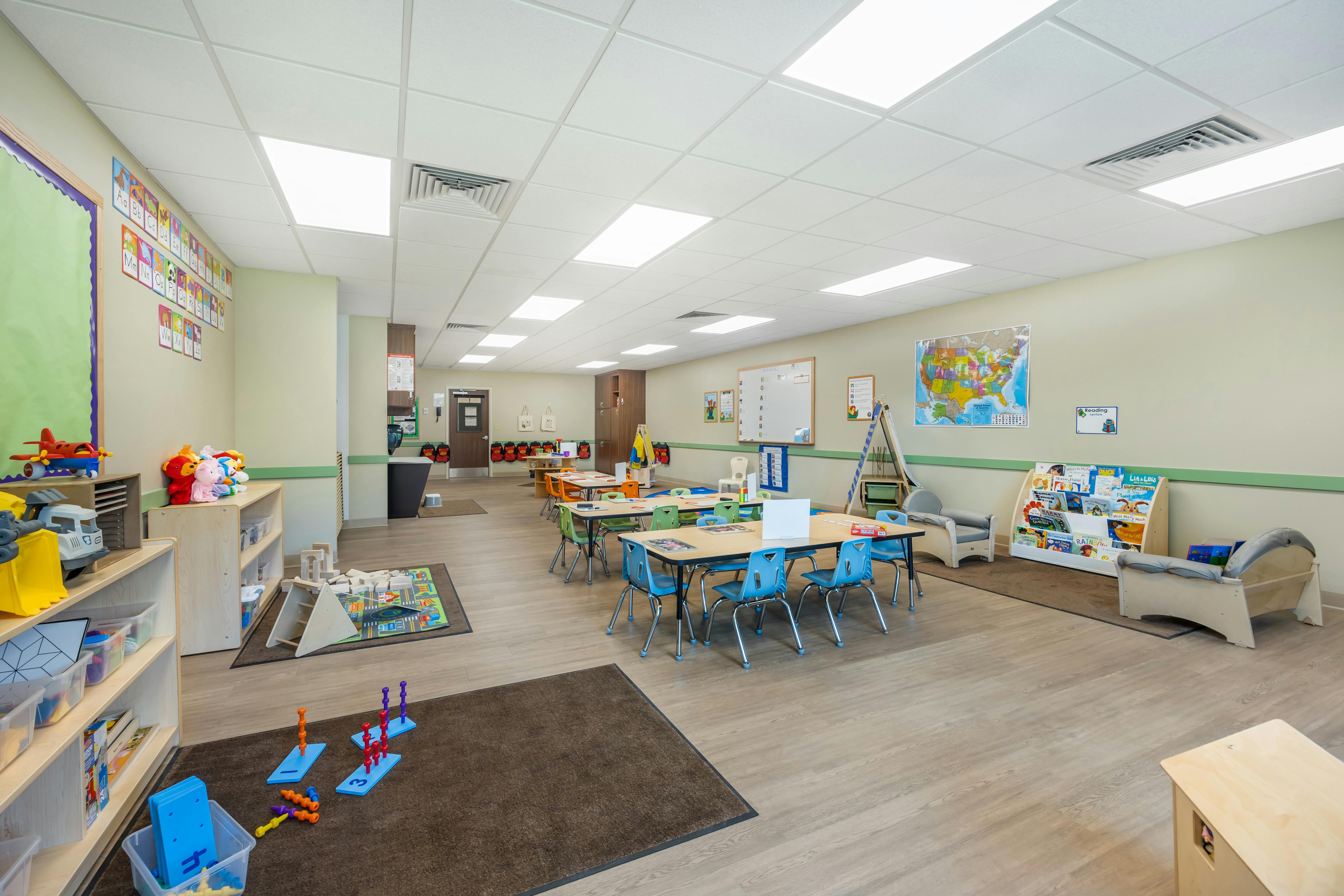 Classroom with small tables and chairs, shelves with toys, and activity rugs