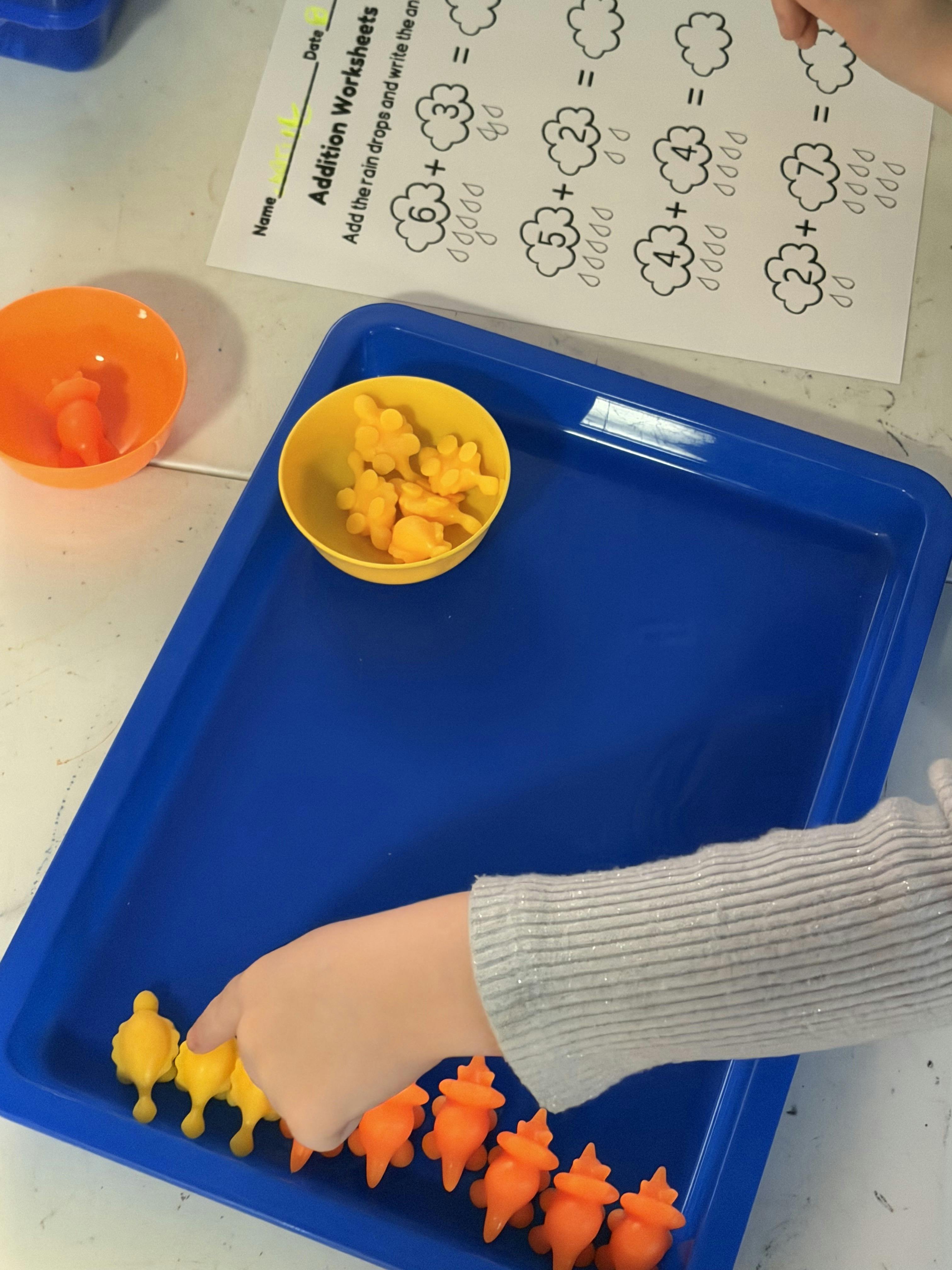 Child counting small plastic figures on a blue tray with an addition worksheet