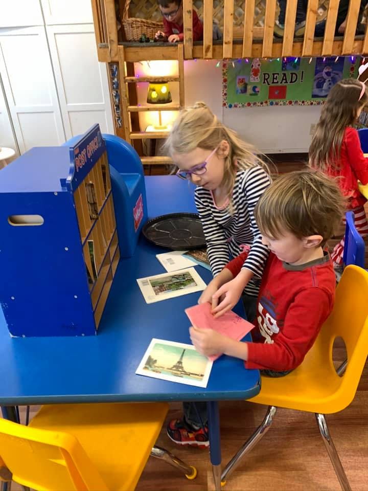 Two children at a blue table with a toy post office and postcards