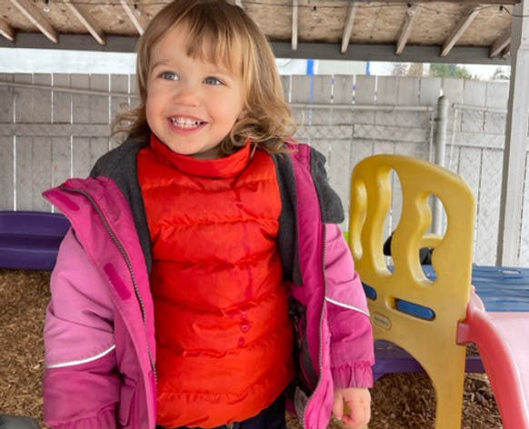 Child smiling while wearing a pink jacket and orange vest outdoors vest