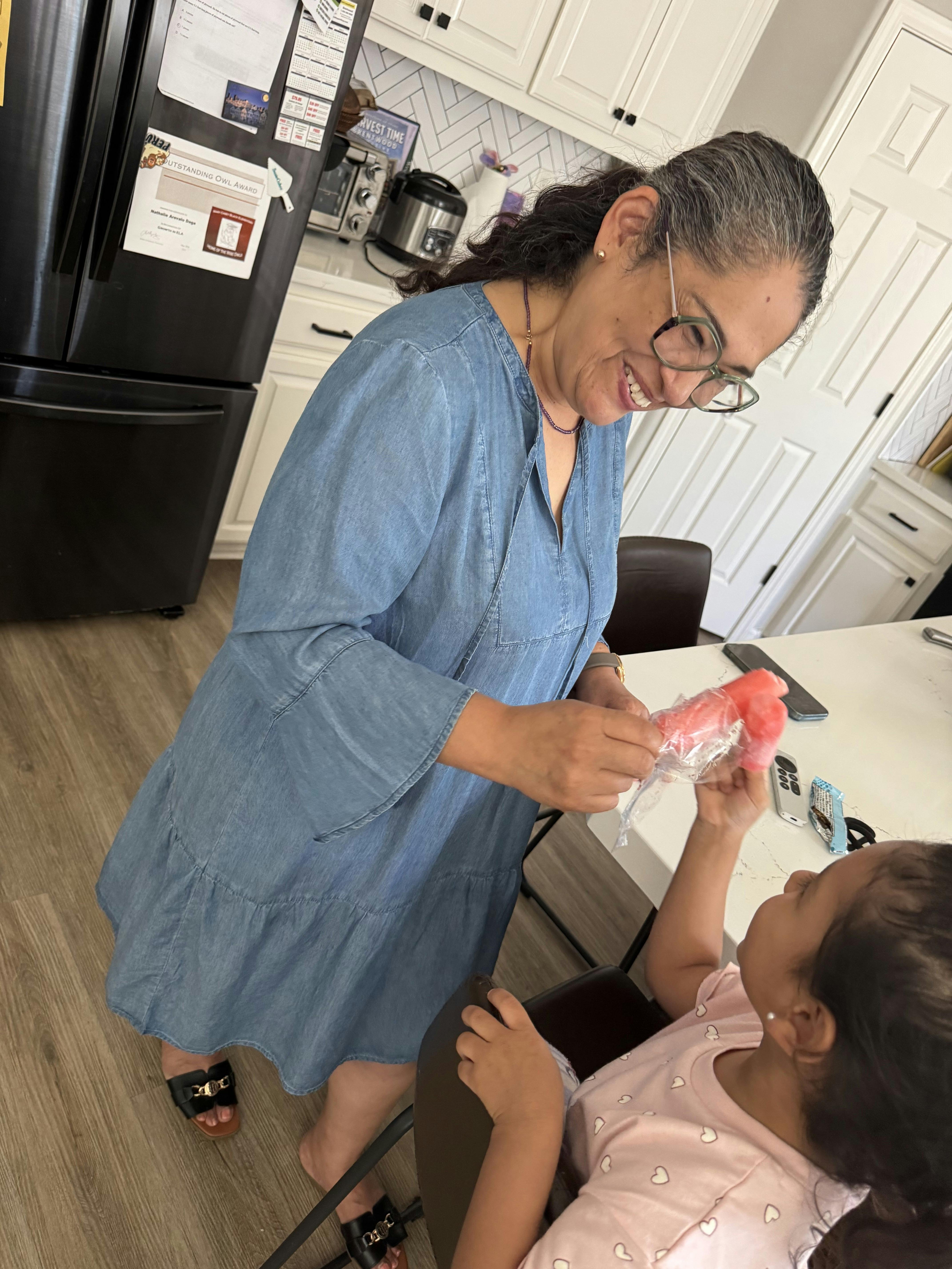 Adult person helping a child unwrap a red frozen treat in a kitchen