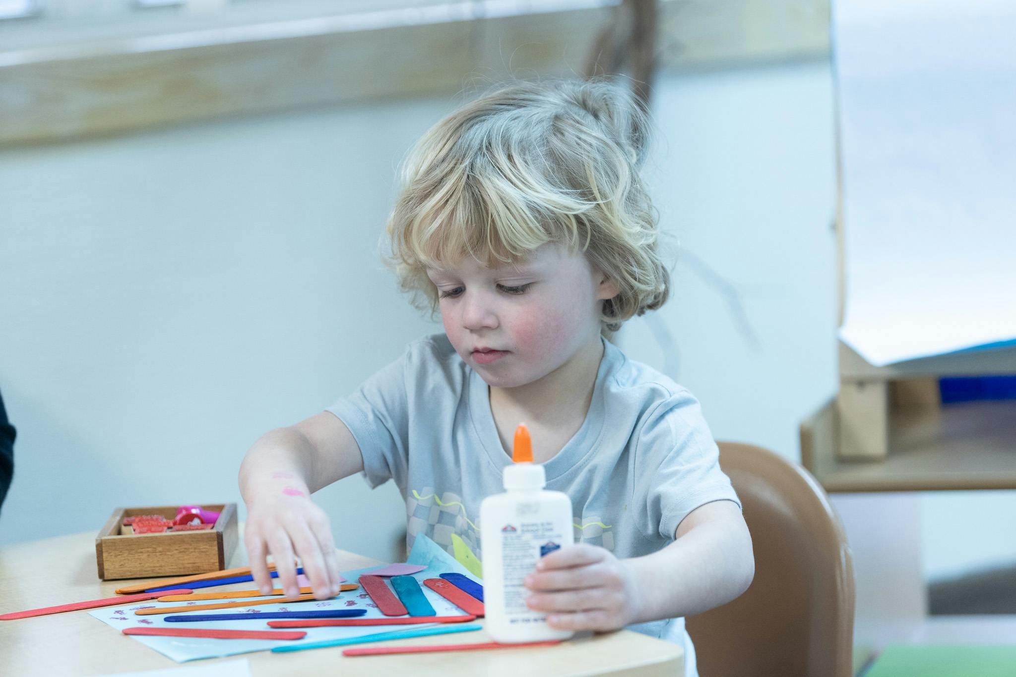 Child holding a bottle of glue and arranging craft sticks on paper
