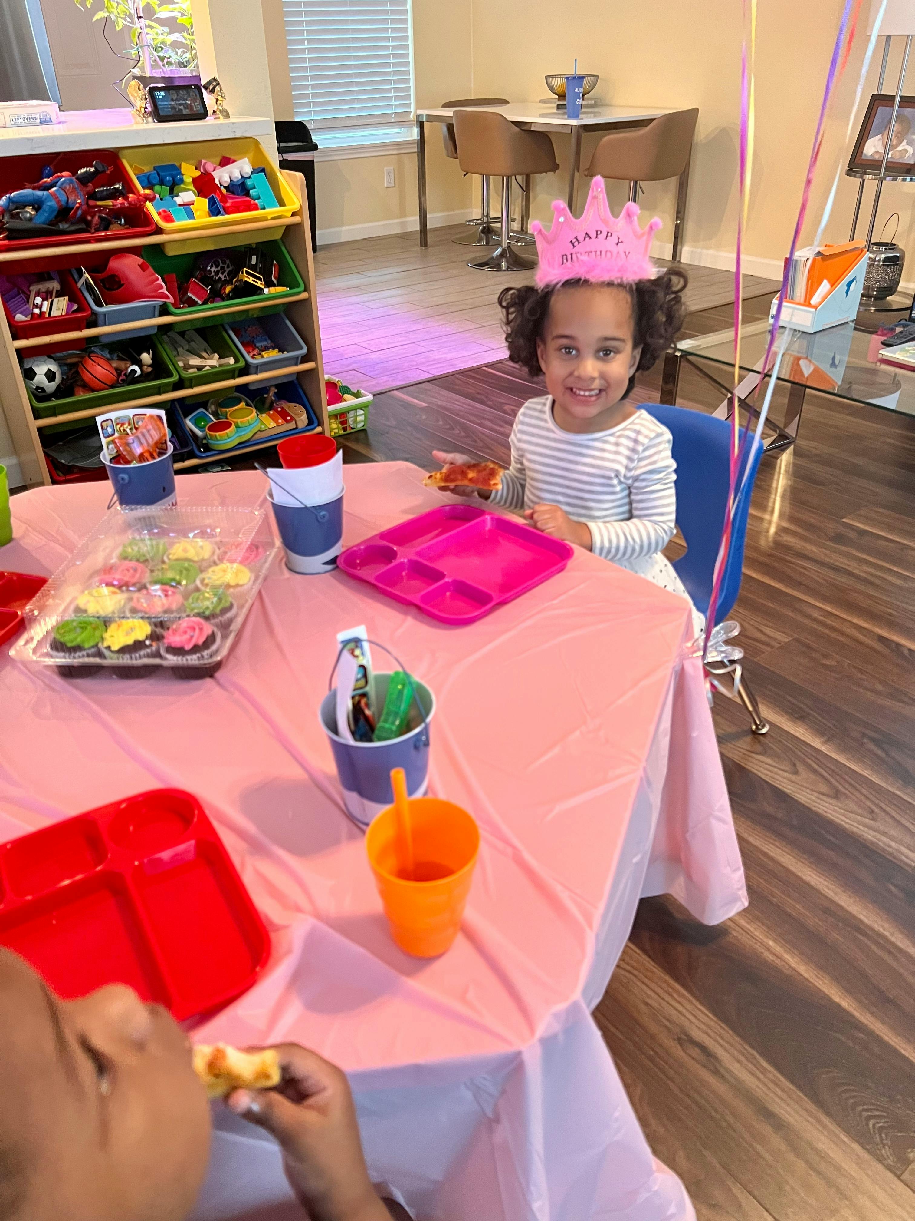 Child wearing a crown sitting at a table with cupcakes and another child eating