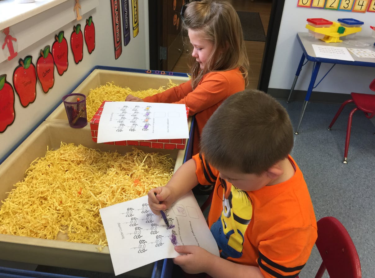 Two children writing at a table with a bin of shredded yellow material