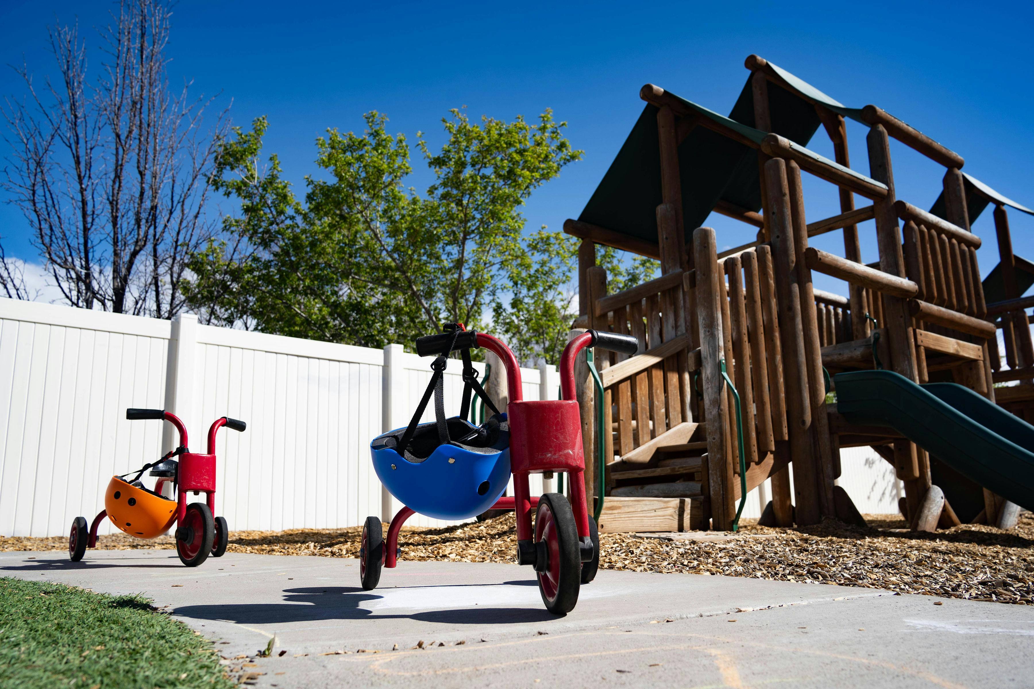 Child-sized tricycles with helmets on a sidewalk next to a playground