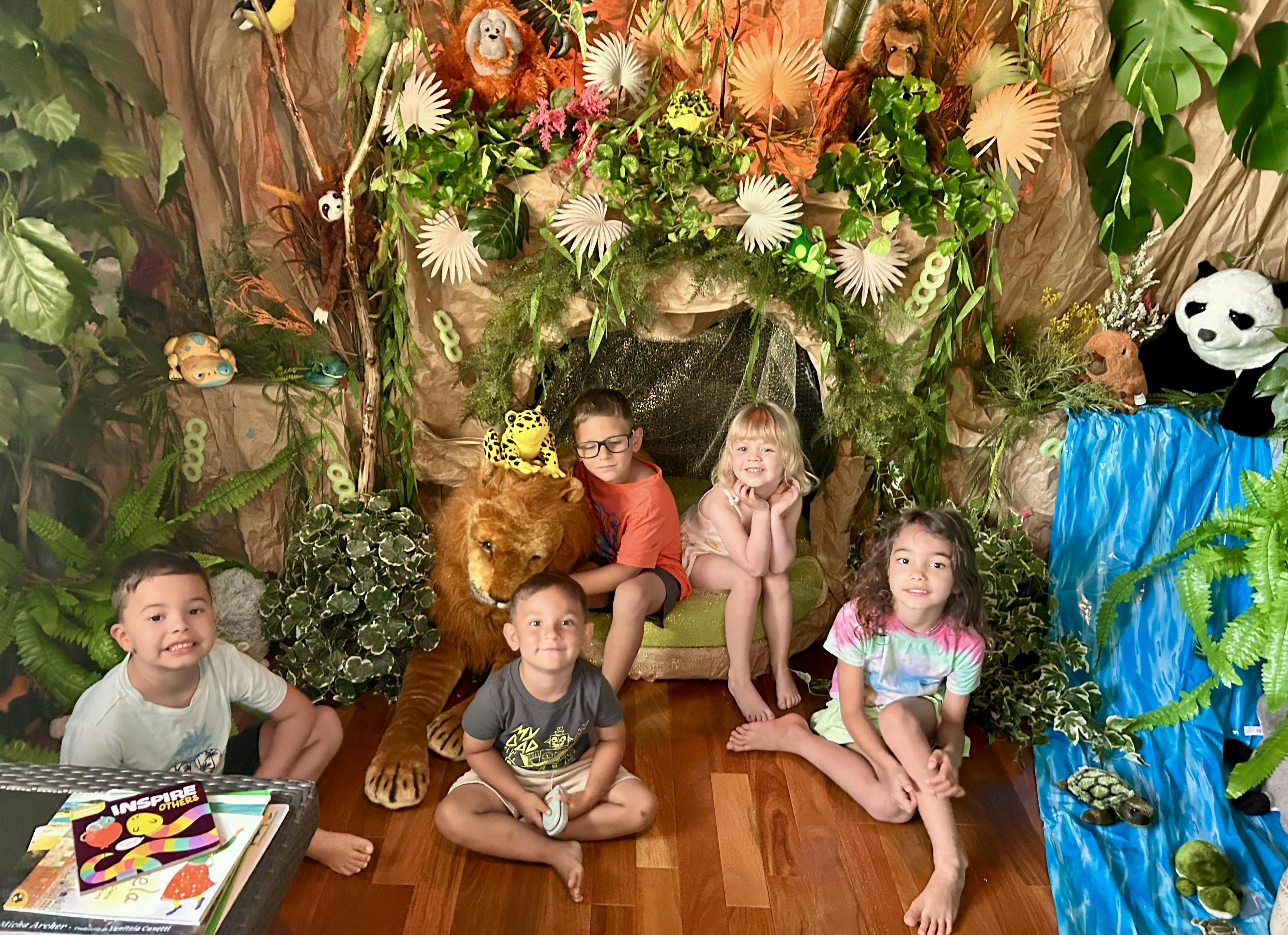 Four children in a jungle-themed room with a stuffed lion, panda, and waterfall