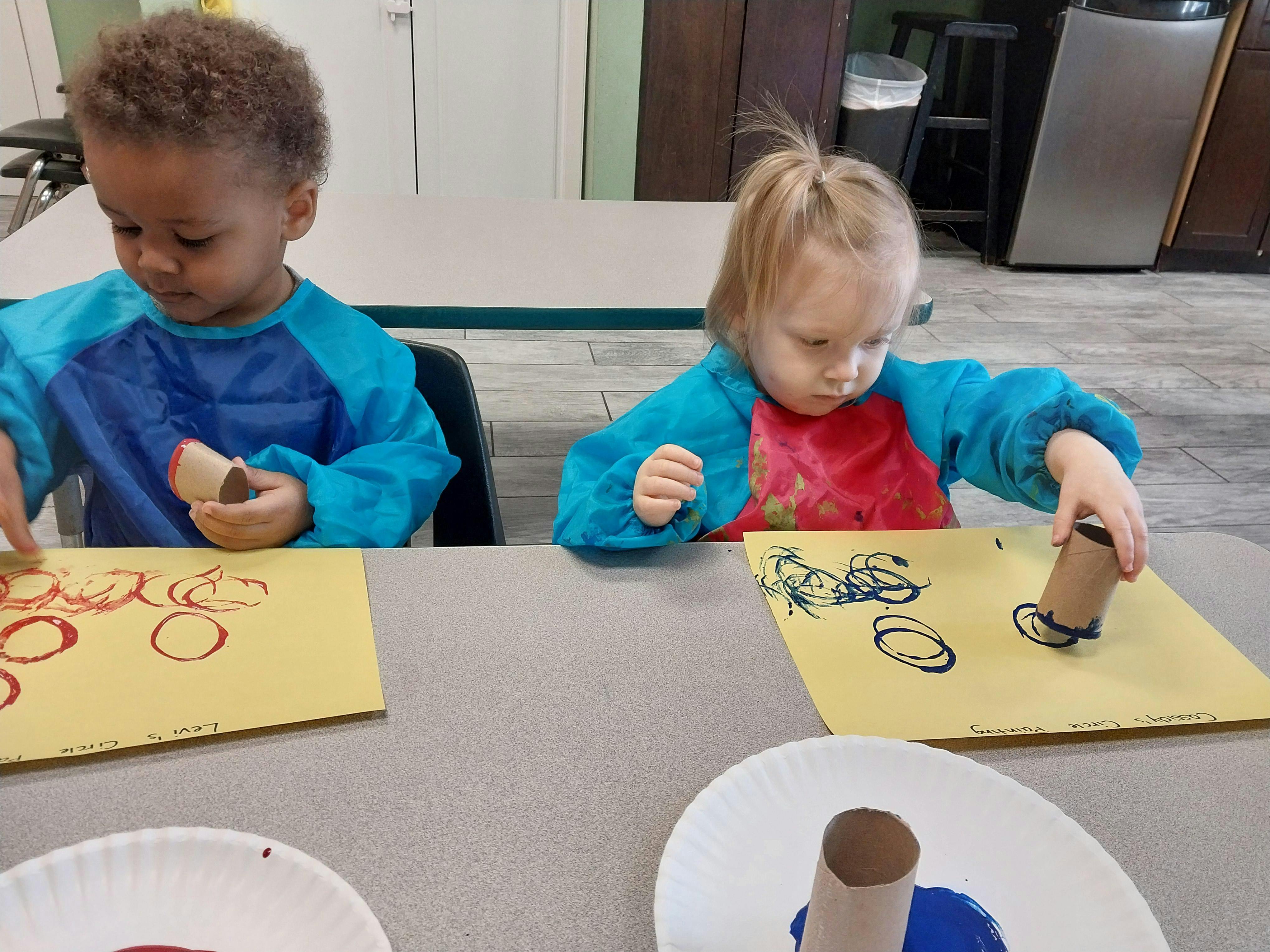 Two children wearing art smocks stamping circles onto paper with cardboard tubes