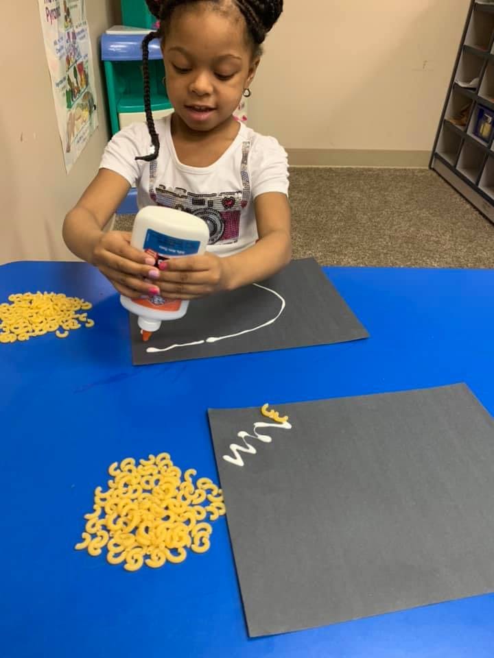 Child squeezing glue onto black paper next to macaroni noodles
