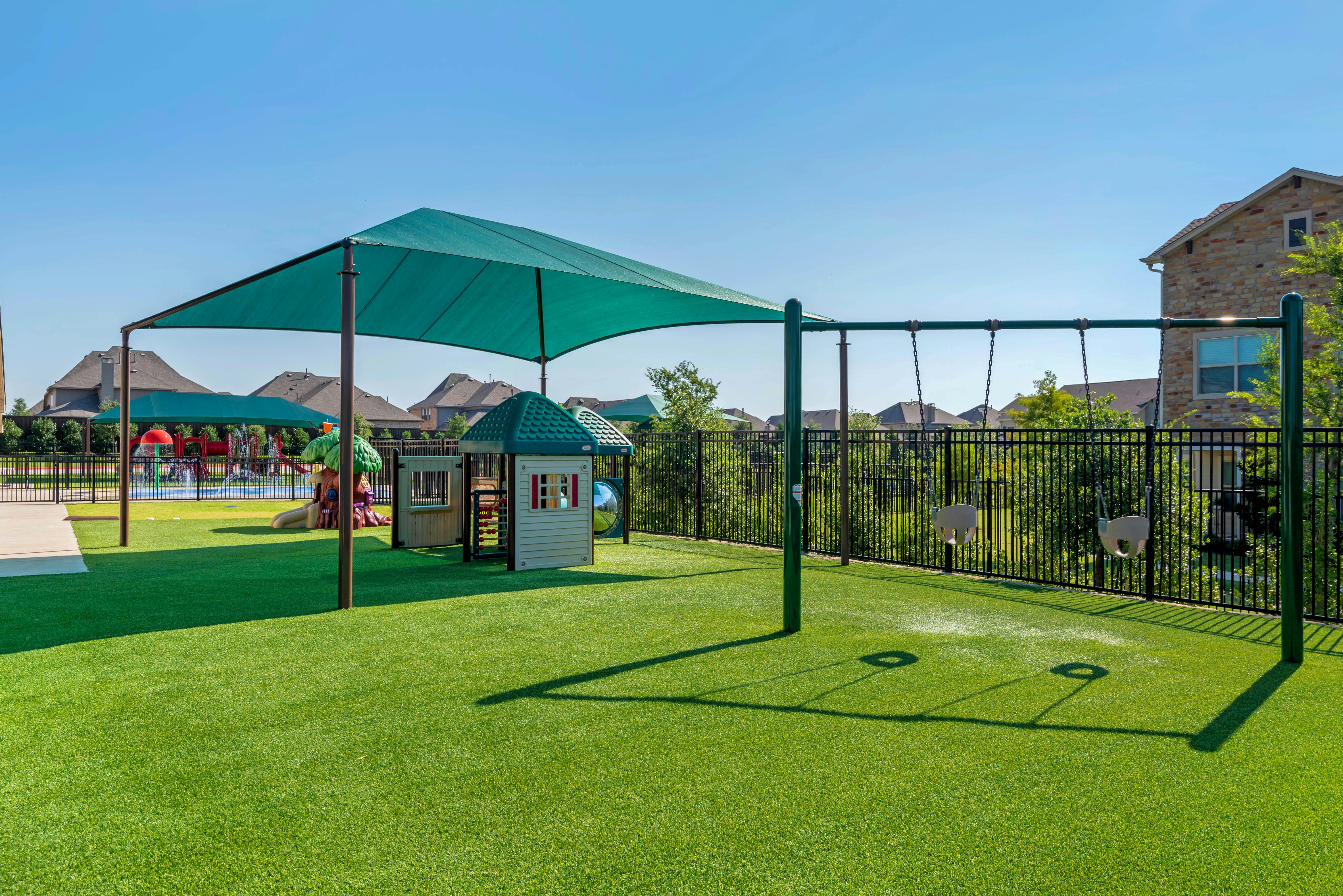 A fenced playground with a green shade, playhouse, and baby swings