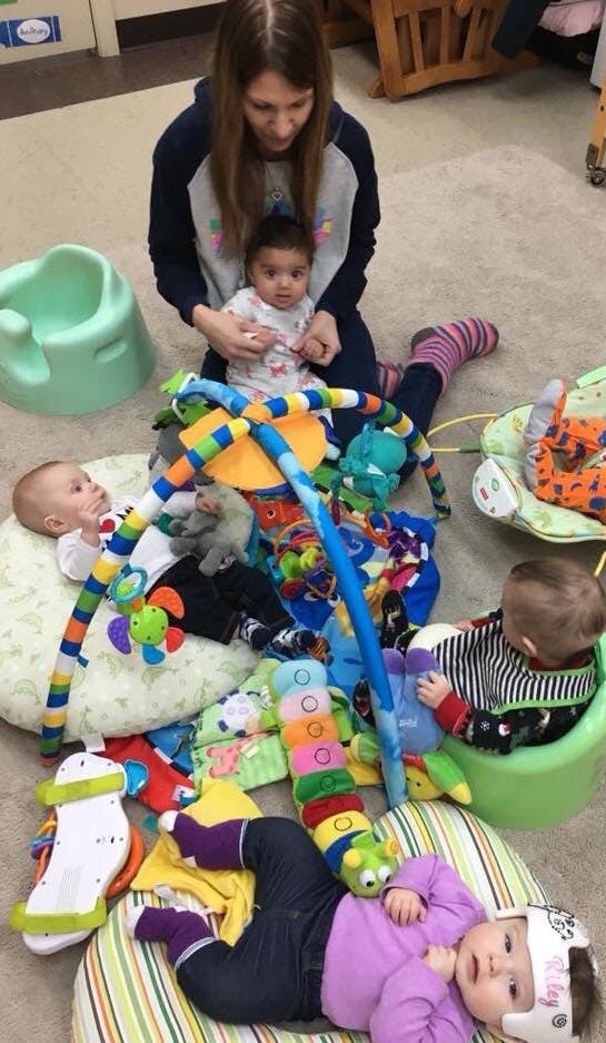 Person kneeling with a child, other children playing with toys on floor with toys