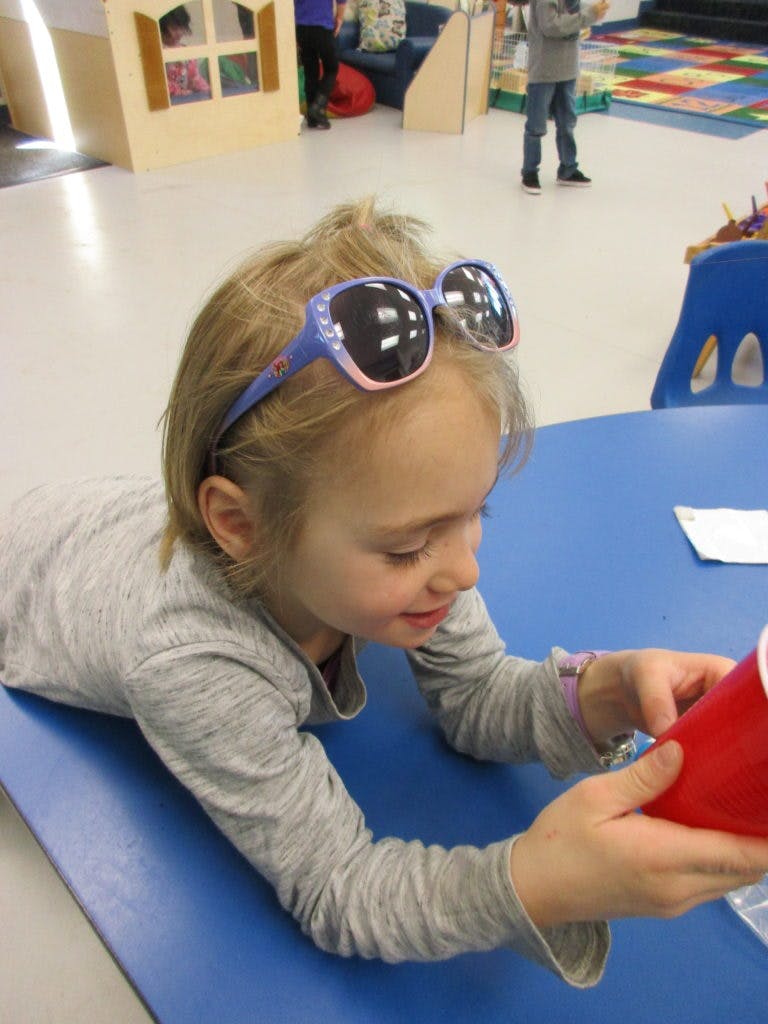 Child with sunglasses on head, looking at a red cup on a blue table