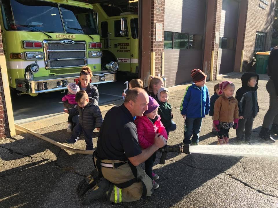Person kneeling with children holding a fire hose, fire truck in background