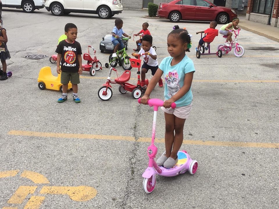Child on a pink scooter with other children on bikes and tricycles