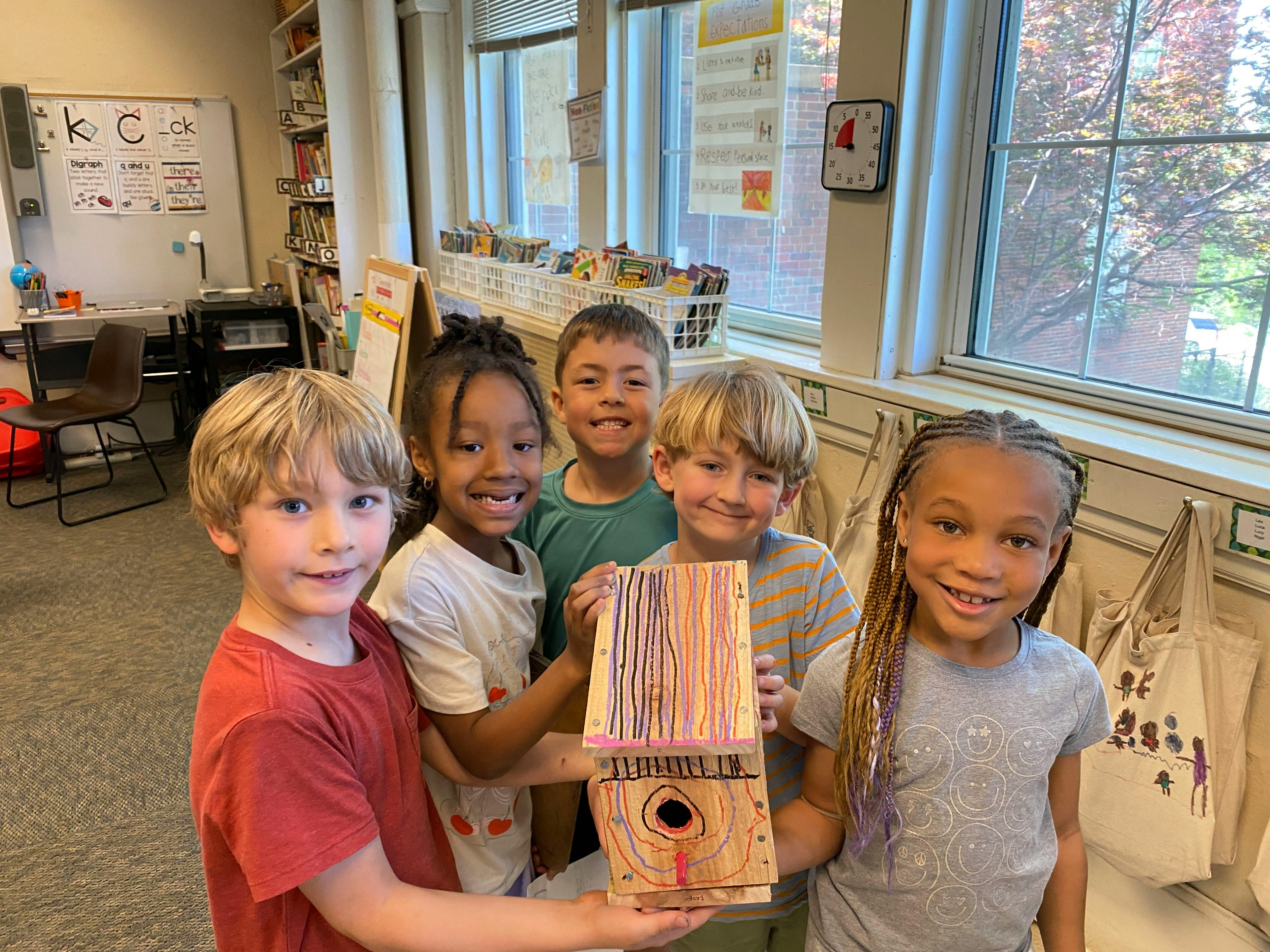 Group of children in a classroom holding a hand-painted wooden birdhouse