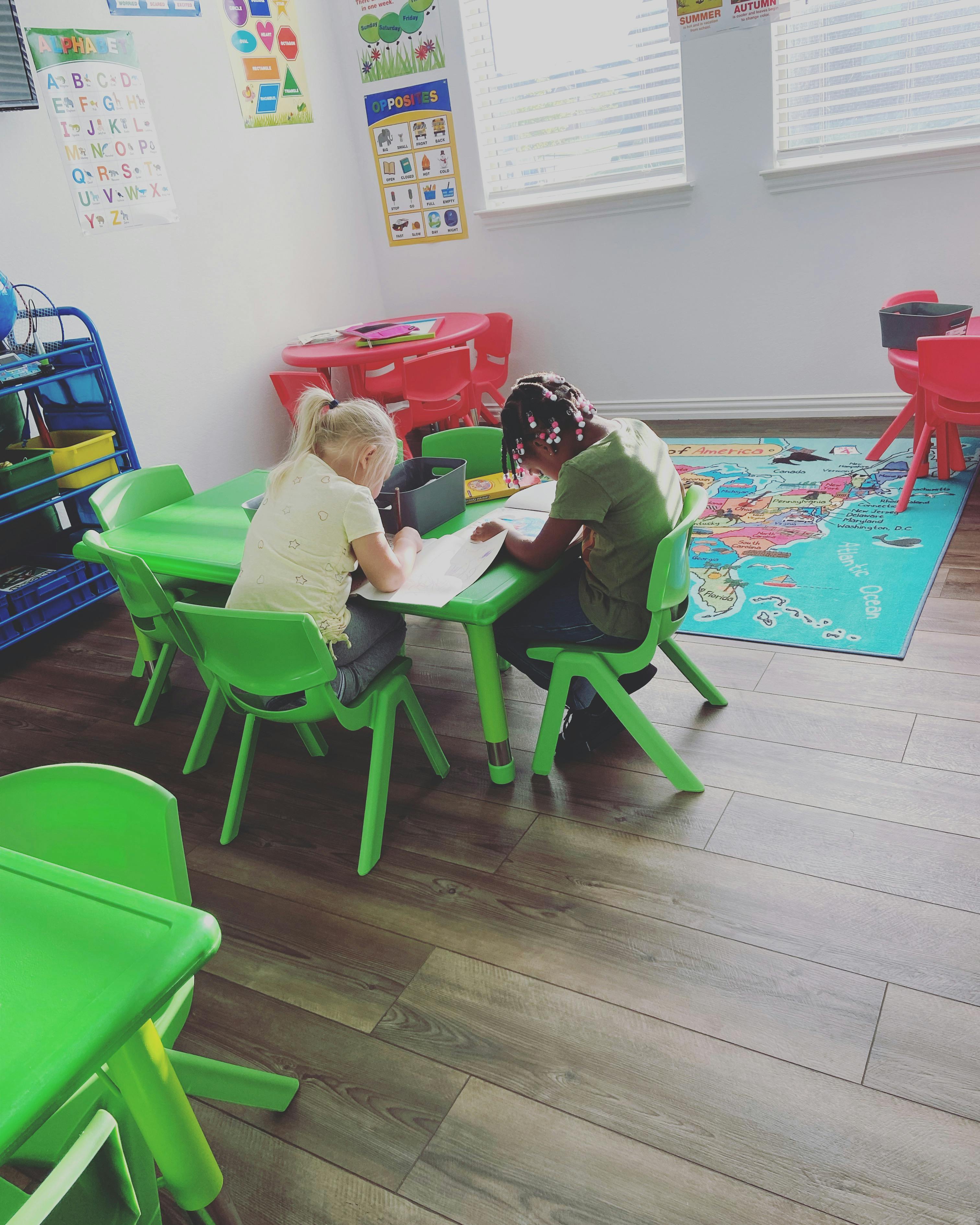 Two children sitting at a green table, looking at papers in a classroom