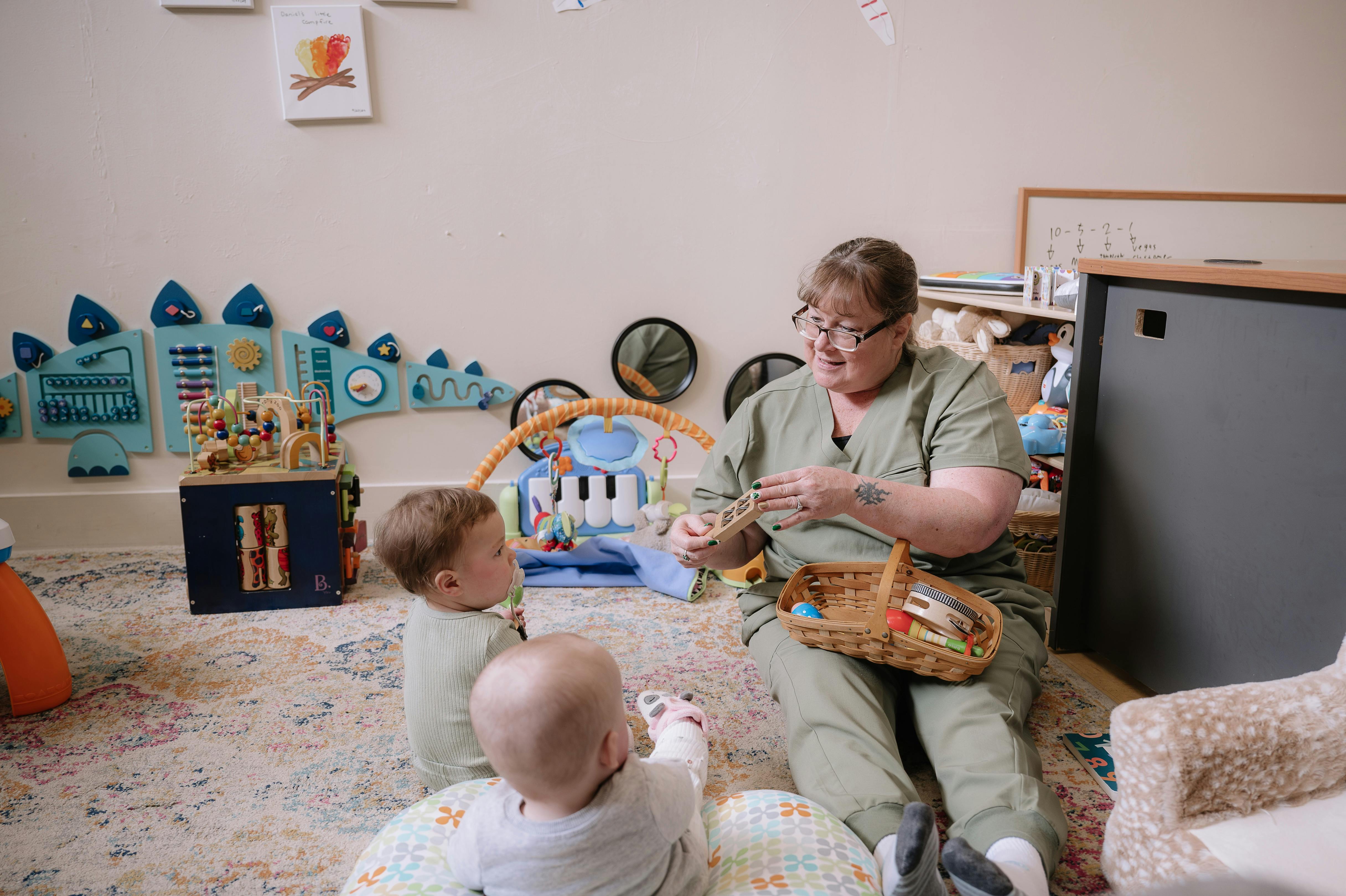 Person sitting on floor with two children, holding a basket of toys