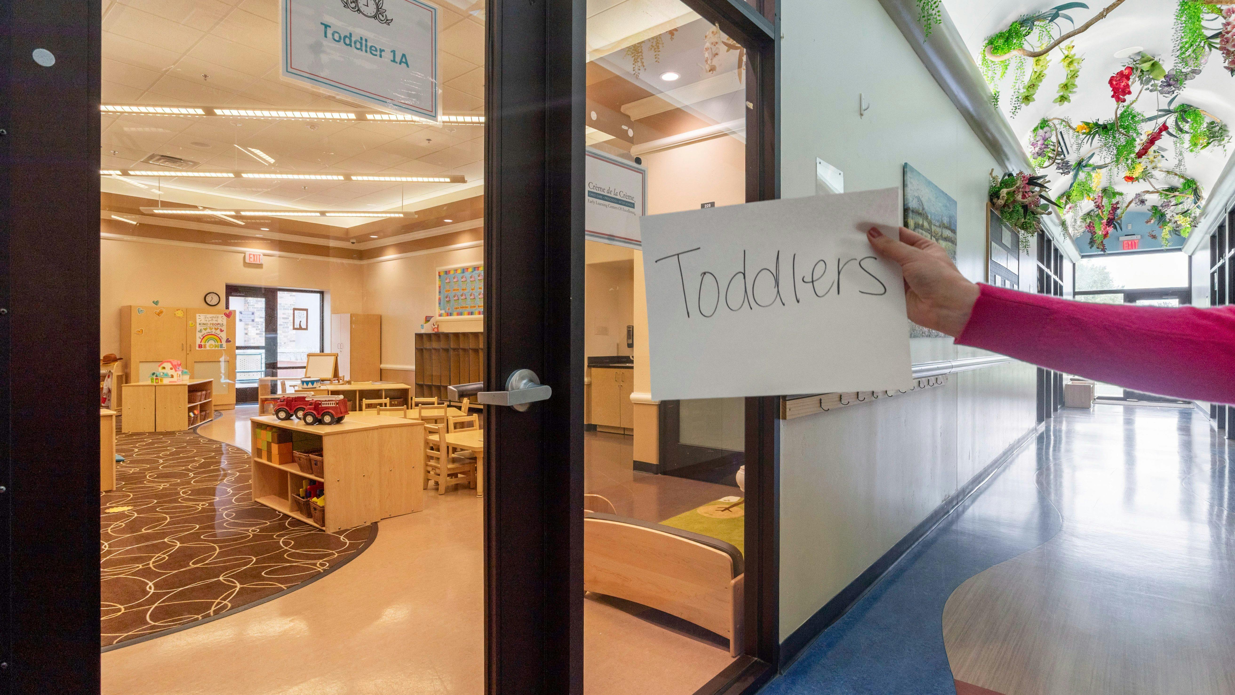 Person holding a "Toddlers" sign next to a glass door into a classroom