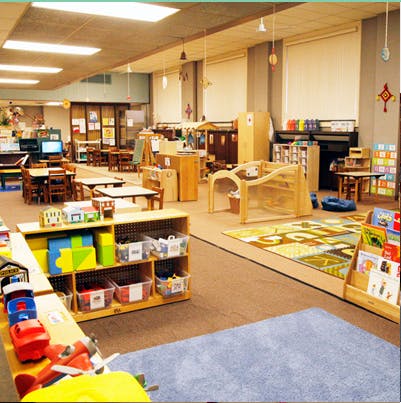 Classroom with small tables and chairs, shelves with toys, and colorful rugs