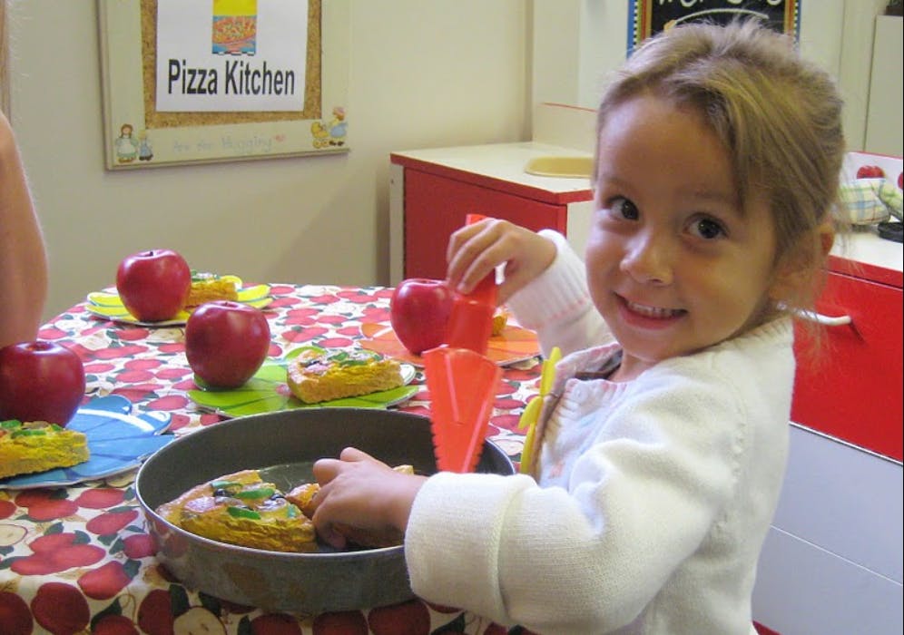 Child playing with toy food at a table with a sign that reads "Pizza Kitchen"