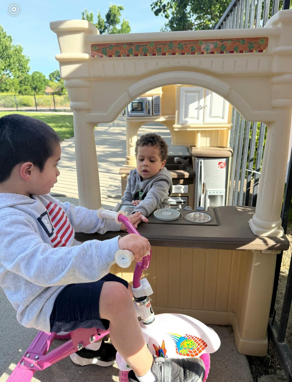 Child on a tricycle looking at another child in a play kitchen