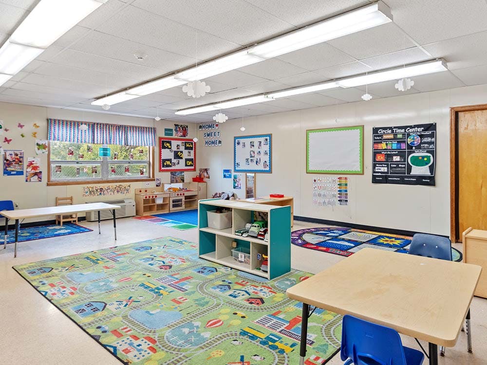 Empty classroom with small tables and chairs, colorful rugs, and learning charts