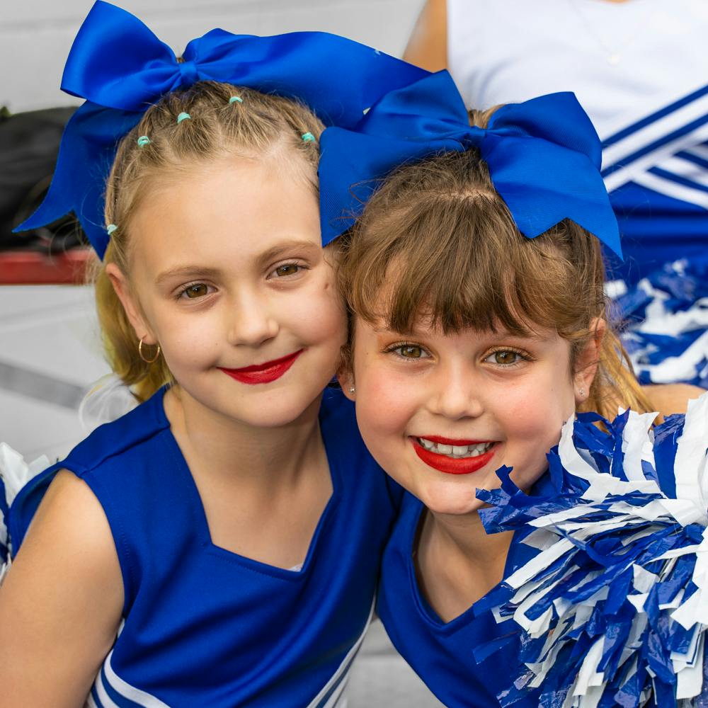 Two children in blue cheerleading outfits with red lipstick and blue bows