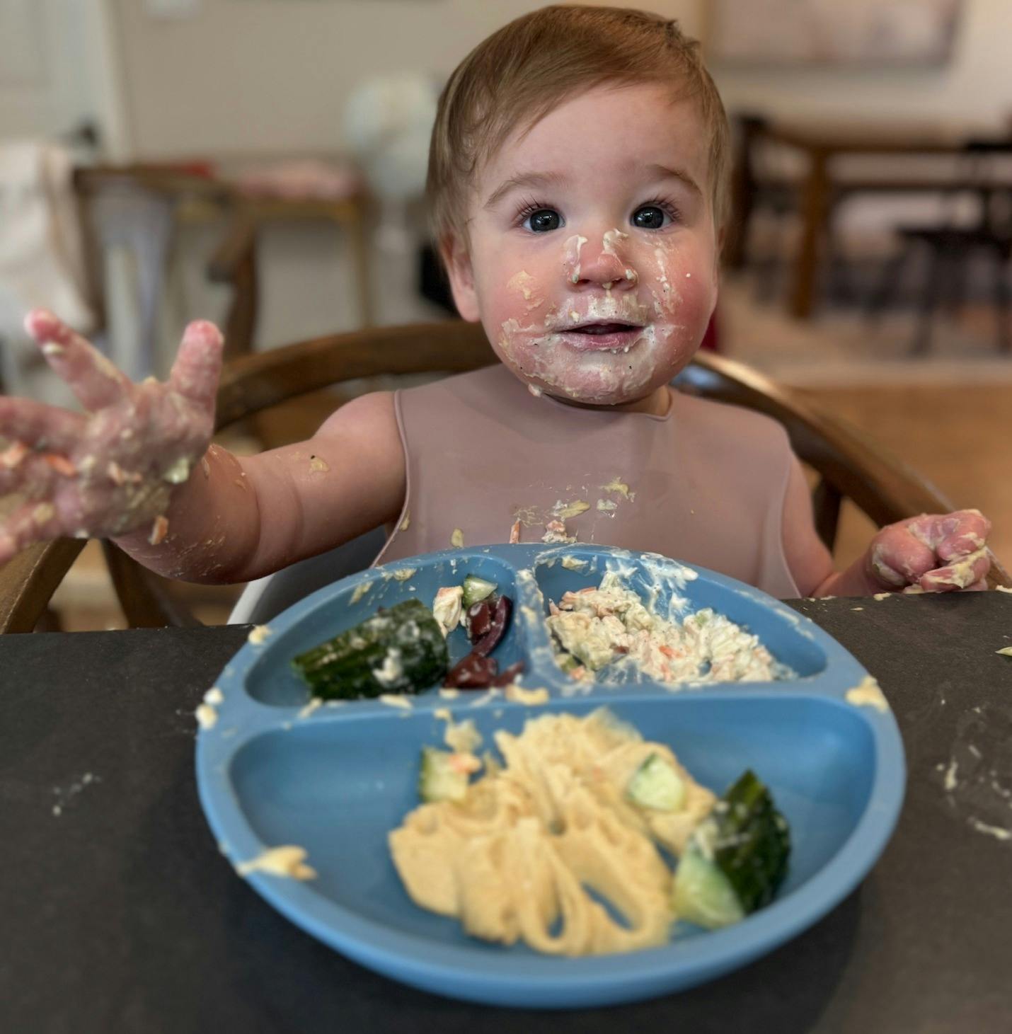 Child with food on face and hands, sitting at a table with a plate of food
