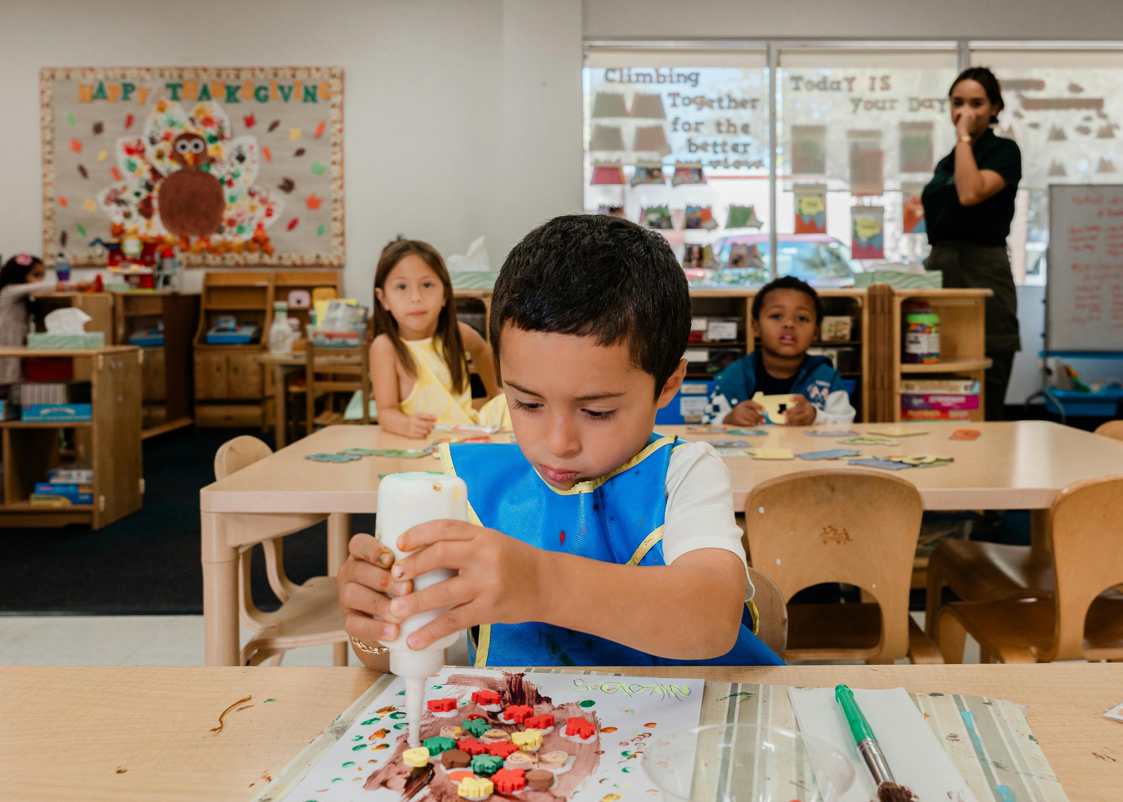 Child in a blue bib squeezing glue onto a craft project in a classroom