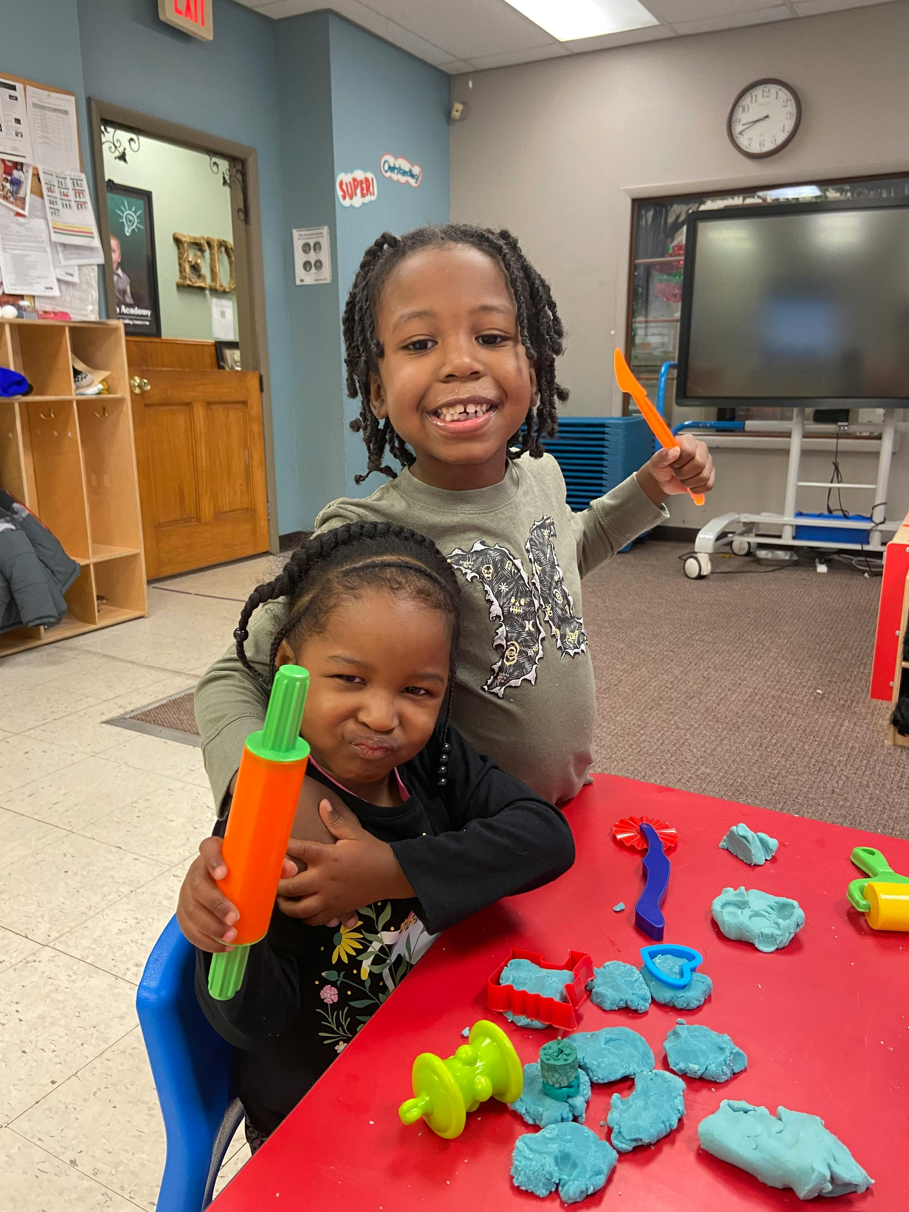 Two children playing with playdough and tools at a red table in a classroom