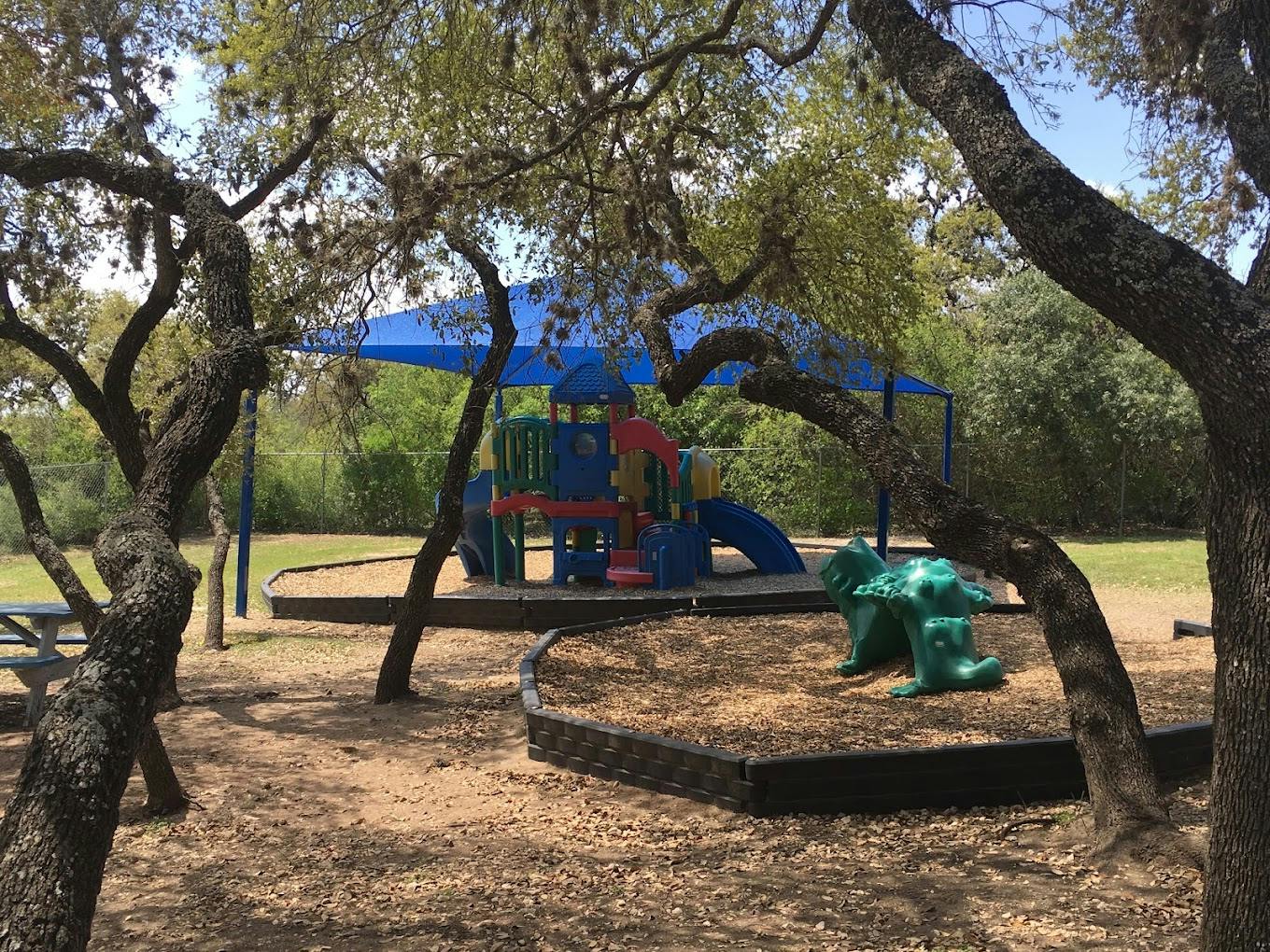 Playground equipment under a blue shade canopy in a wooded area