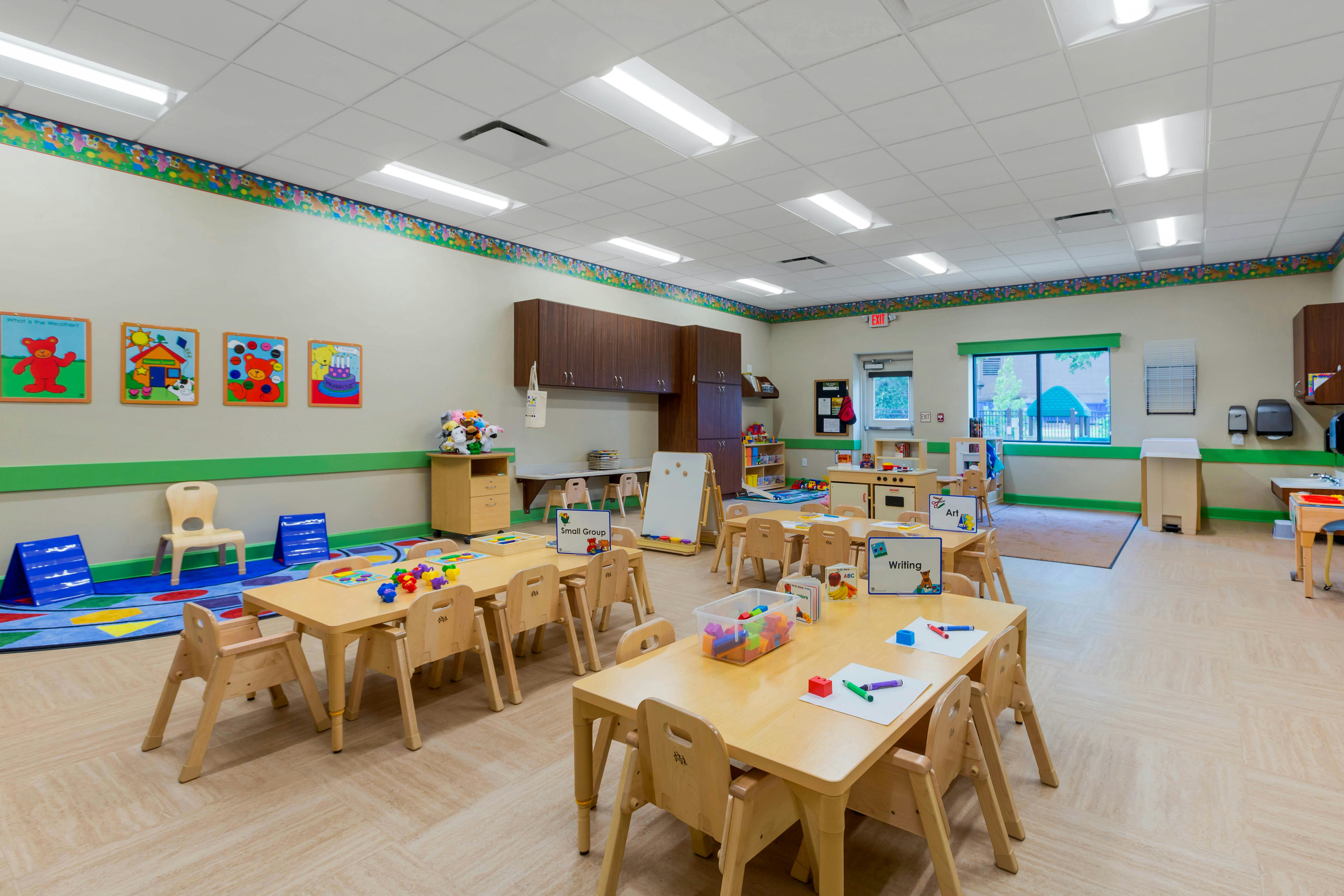 Classroom with small wooden tables and chairs, toys on tables, and colorful rugs