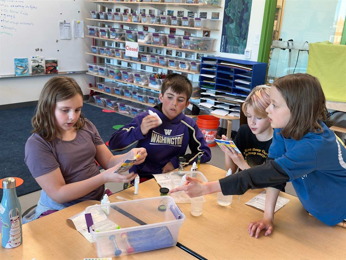 Four children at a table during a science experiment, two looking at instructions