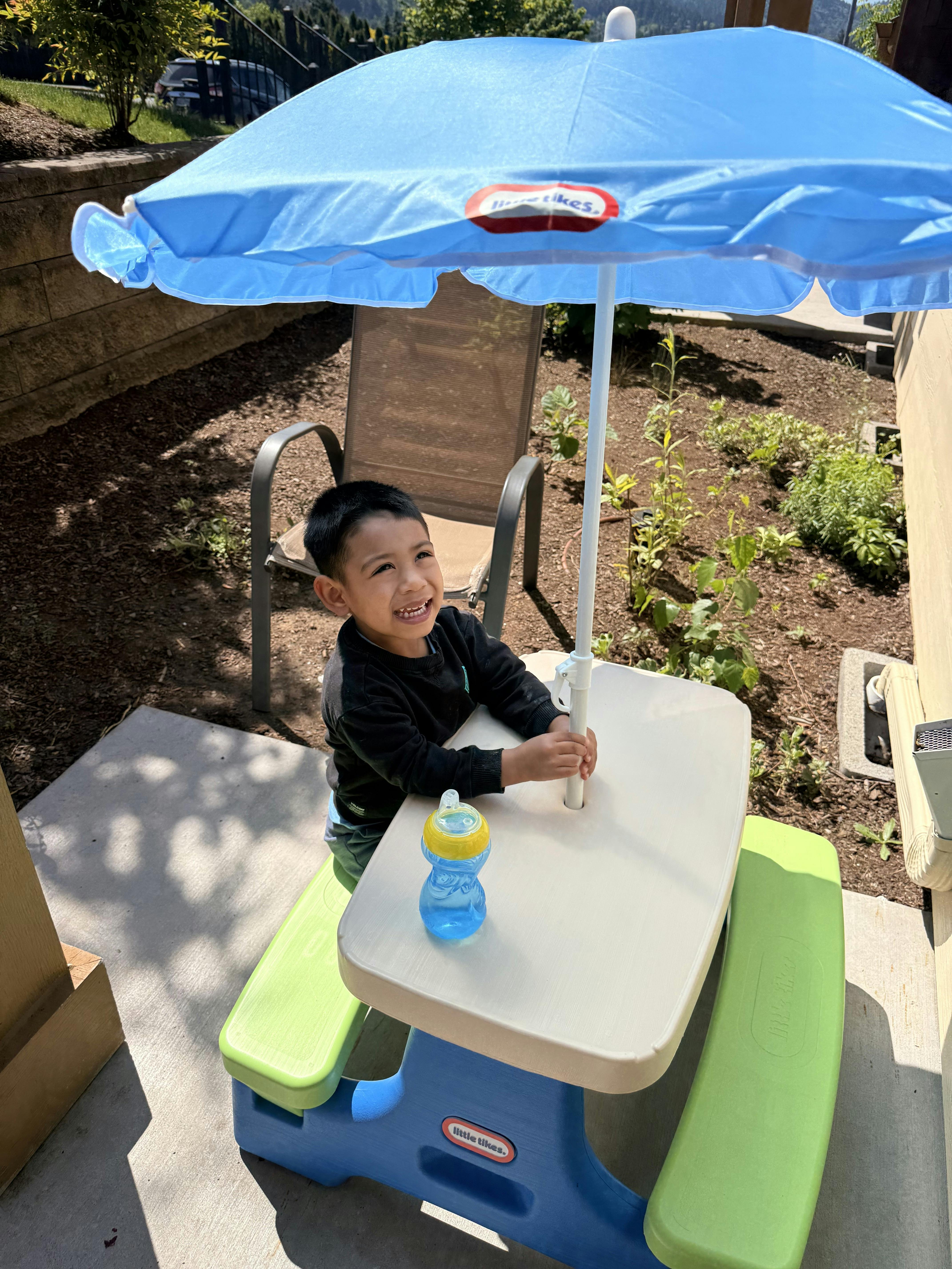 Child sitting at a small picnic table with an umbrella outdoors