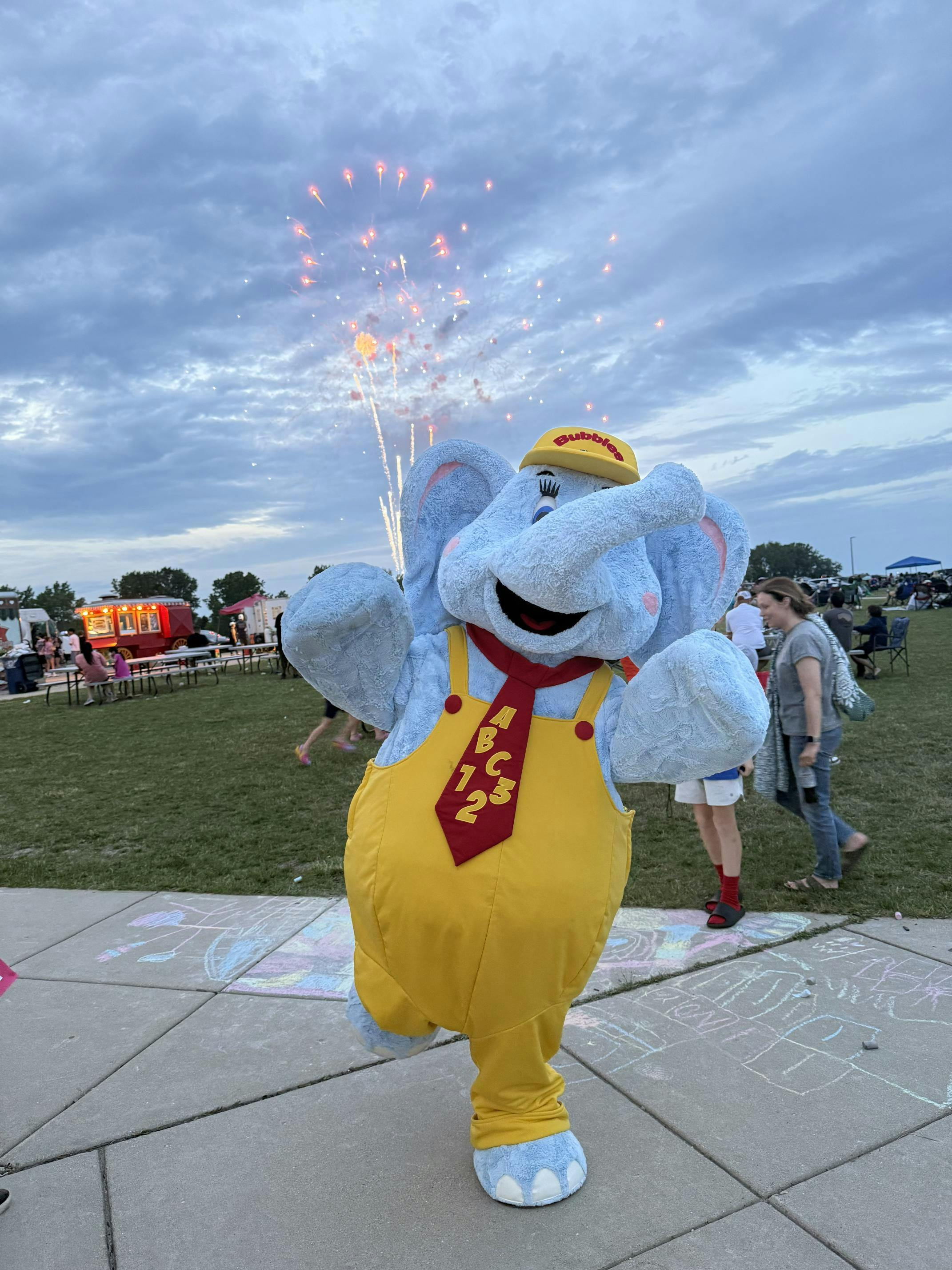 Elephant mascot in yellow overalls and red tie with fireworks in background
