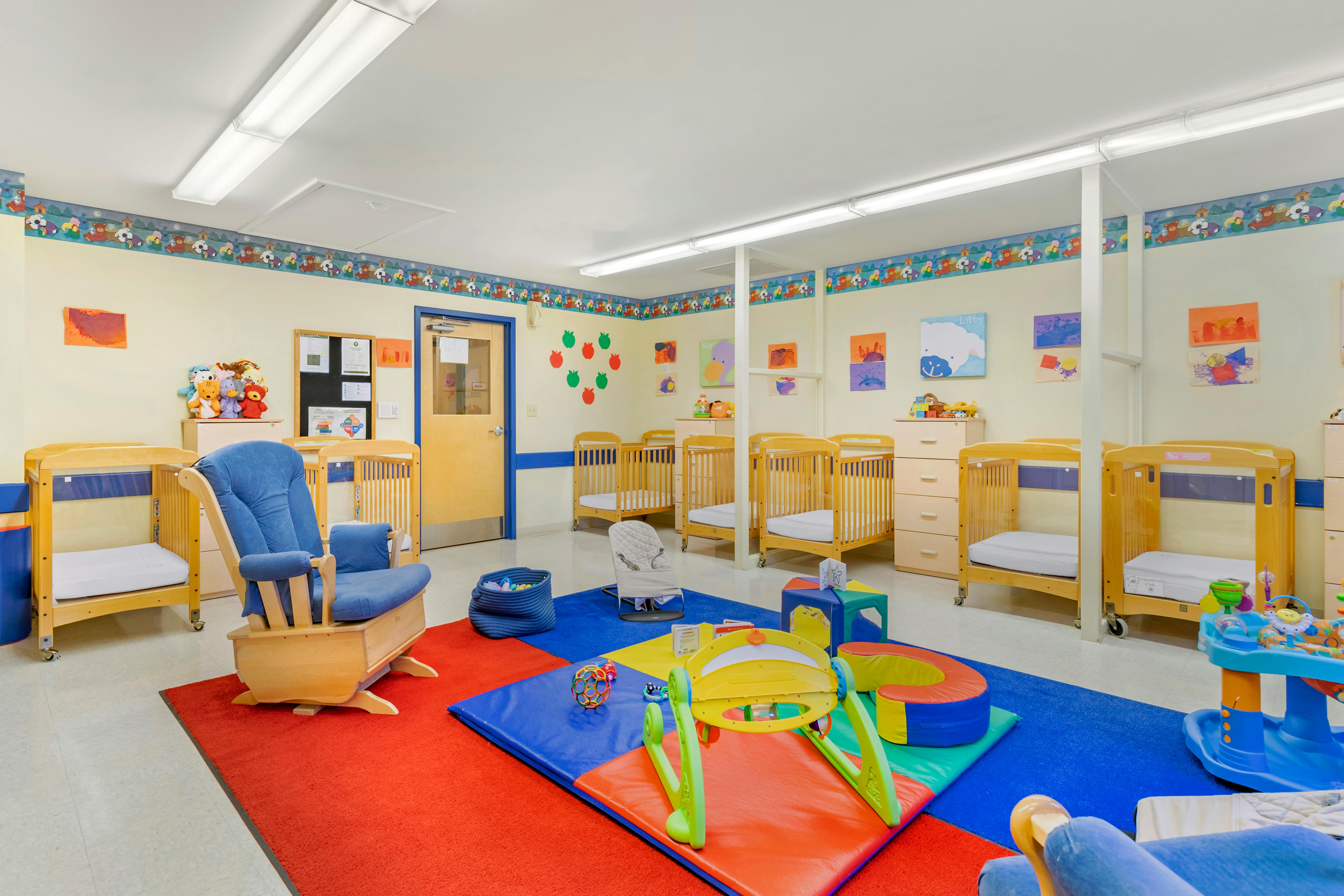 A classroom with several cribs, a rocking chair, and playmats with infant toys