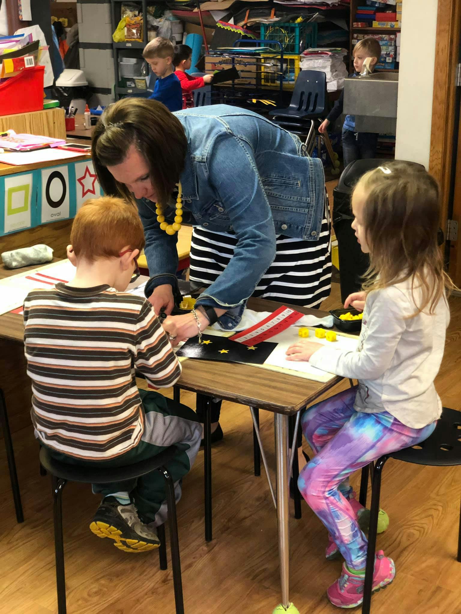 Person helping two children with an art project at a table in a classroom