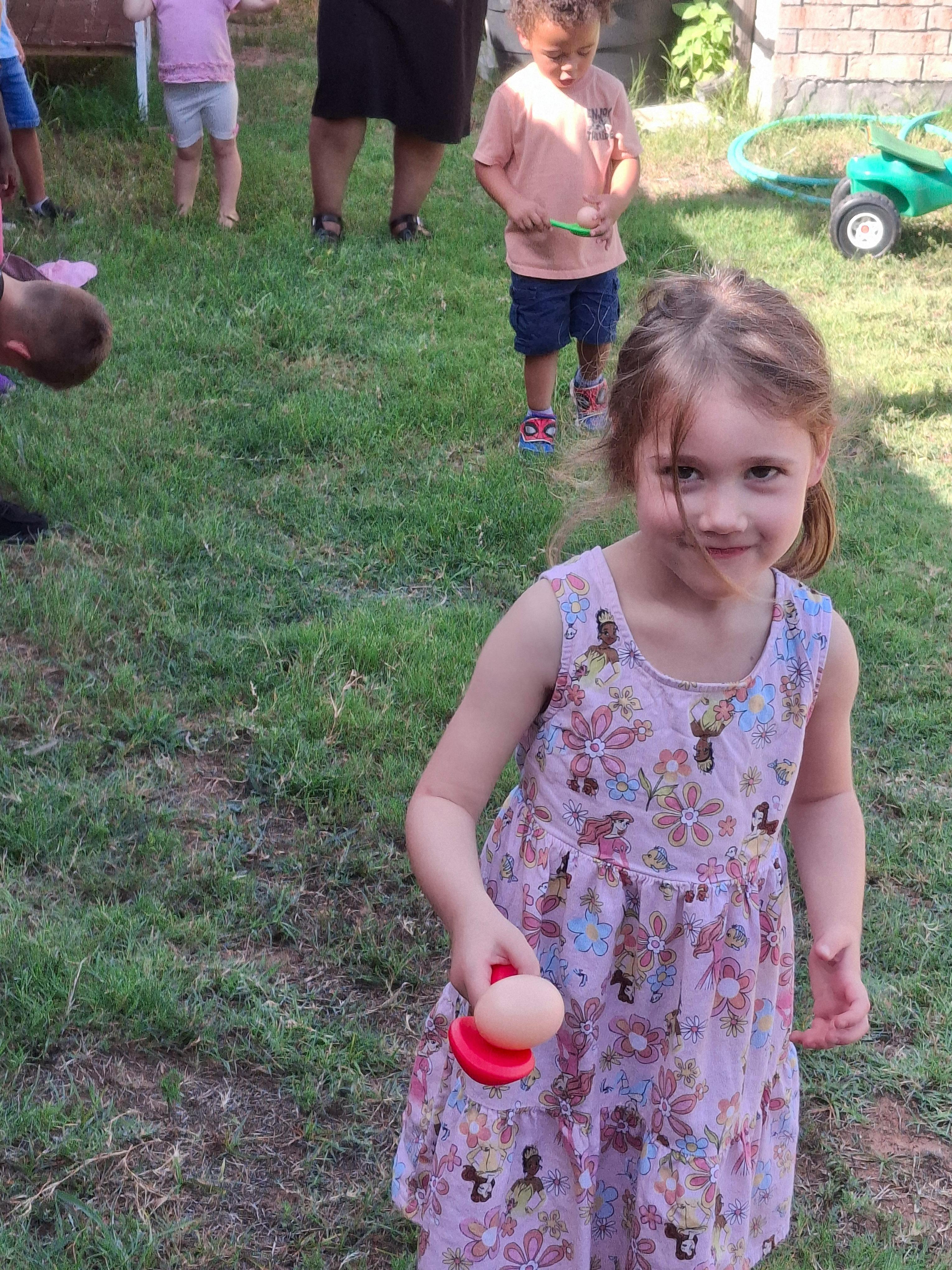 Child in a patterned dress holding a toy with an egg on a lawn