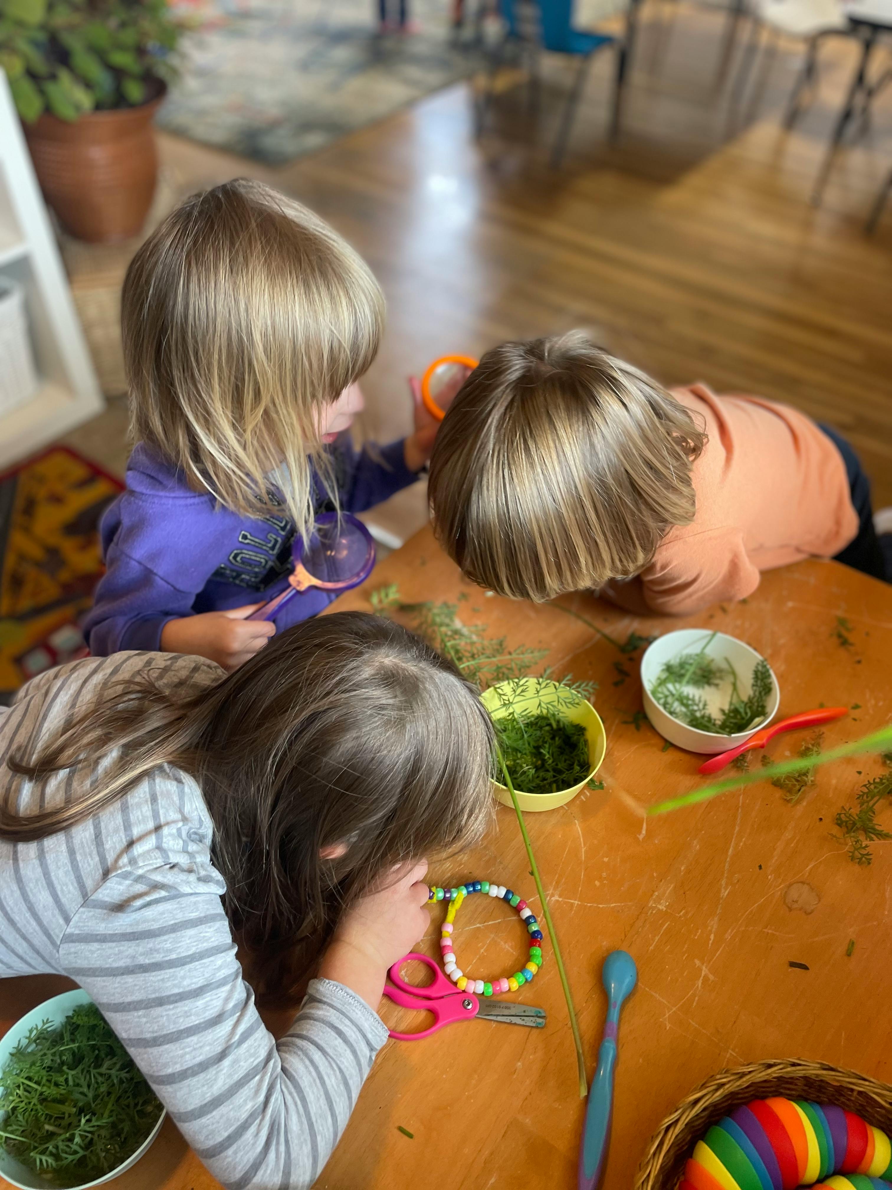 Three children playing with green plant material and craft supplies at a table
