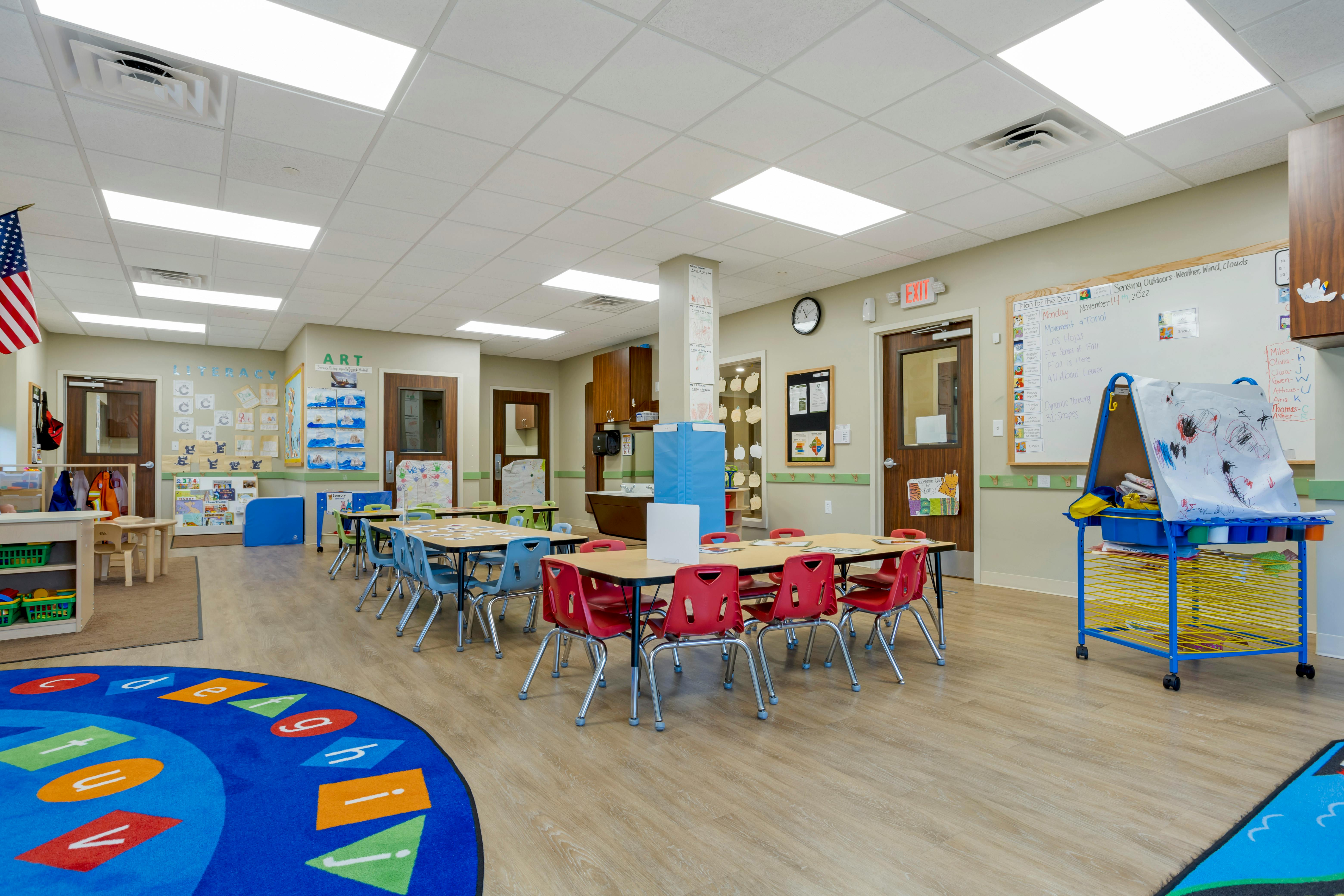 Classroom with small tables and chairs, a blue rug with letters, and whiteboards