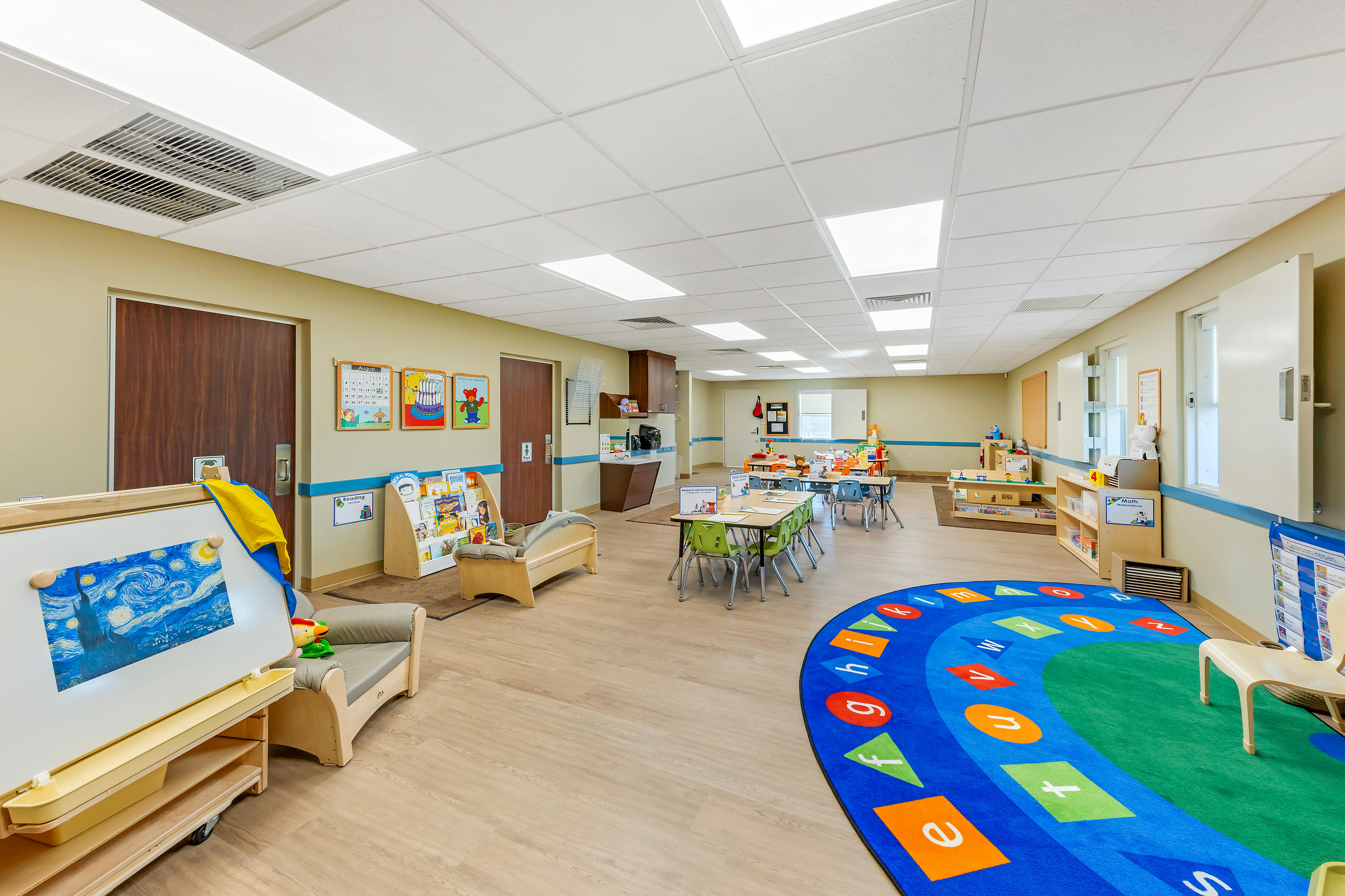 Classroom with small tables and chairs, a play area, and a colorful alphabet rug