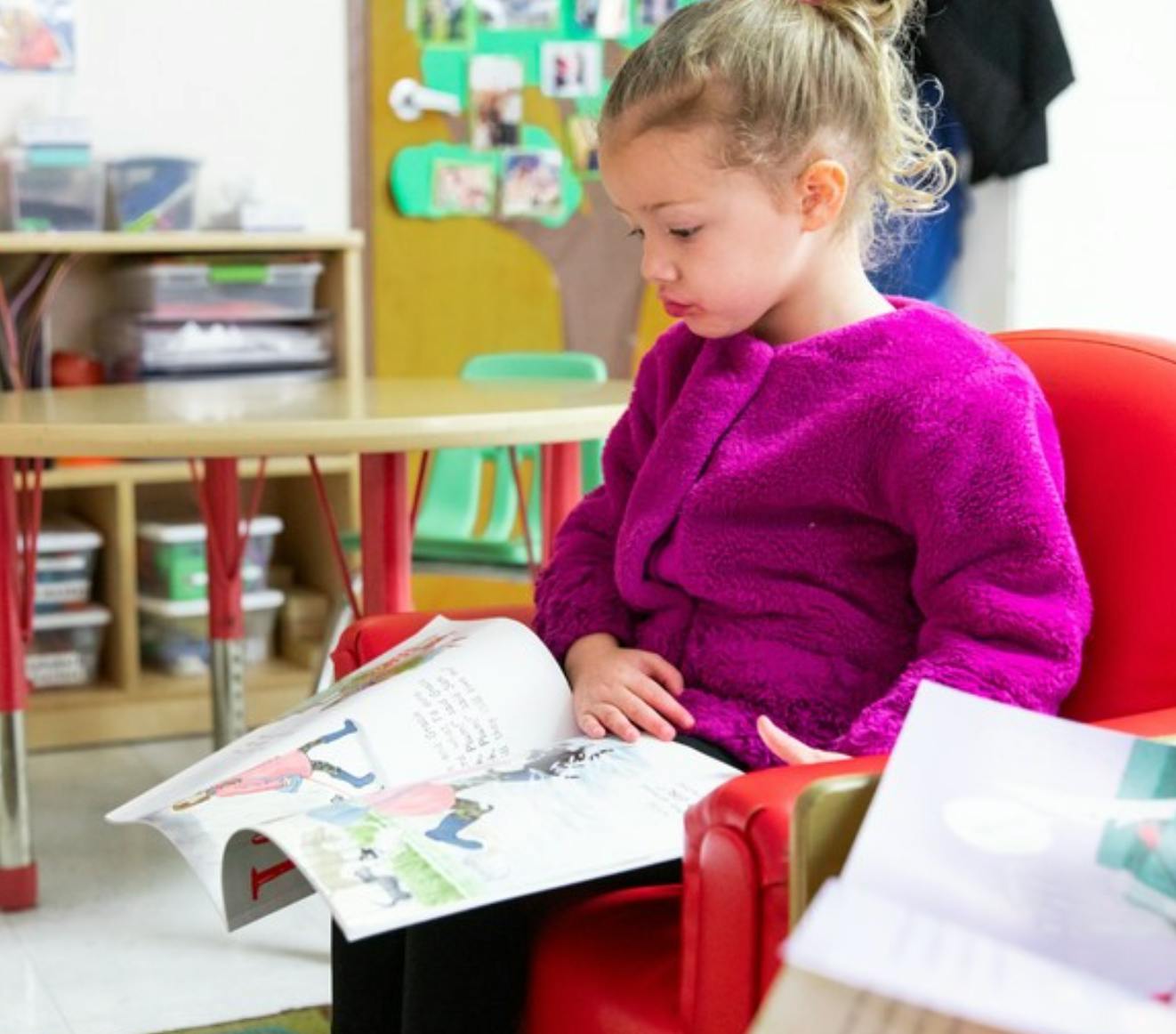 Child in a purple sweater sitting in a red chair reading a book