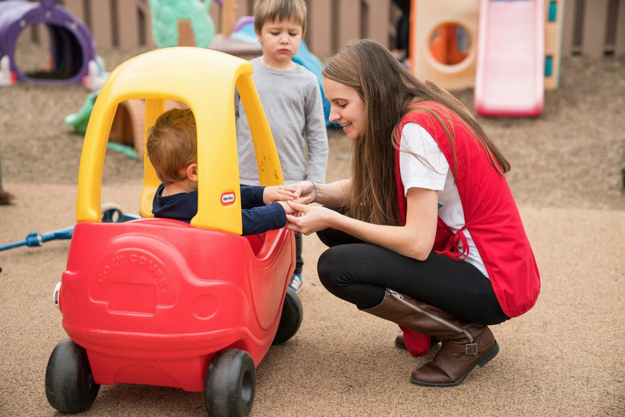 Person crouching next to a child in a toy car, another child standing nearby