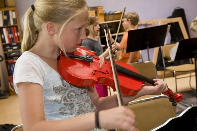 Child playing a red violin in a classroom with other children