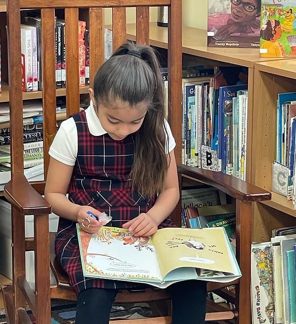 Child sitting in a wooden rocking chair, reading a book in a library