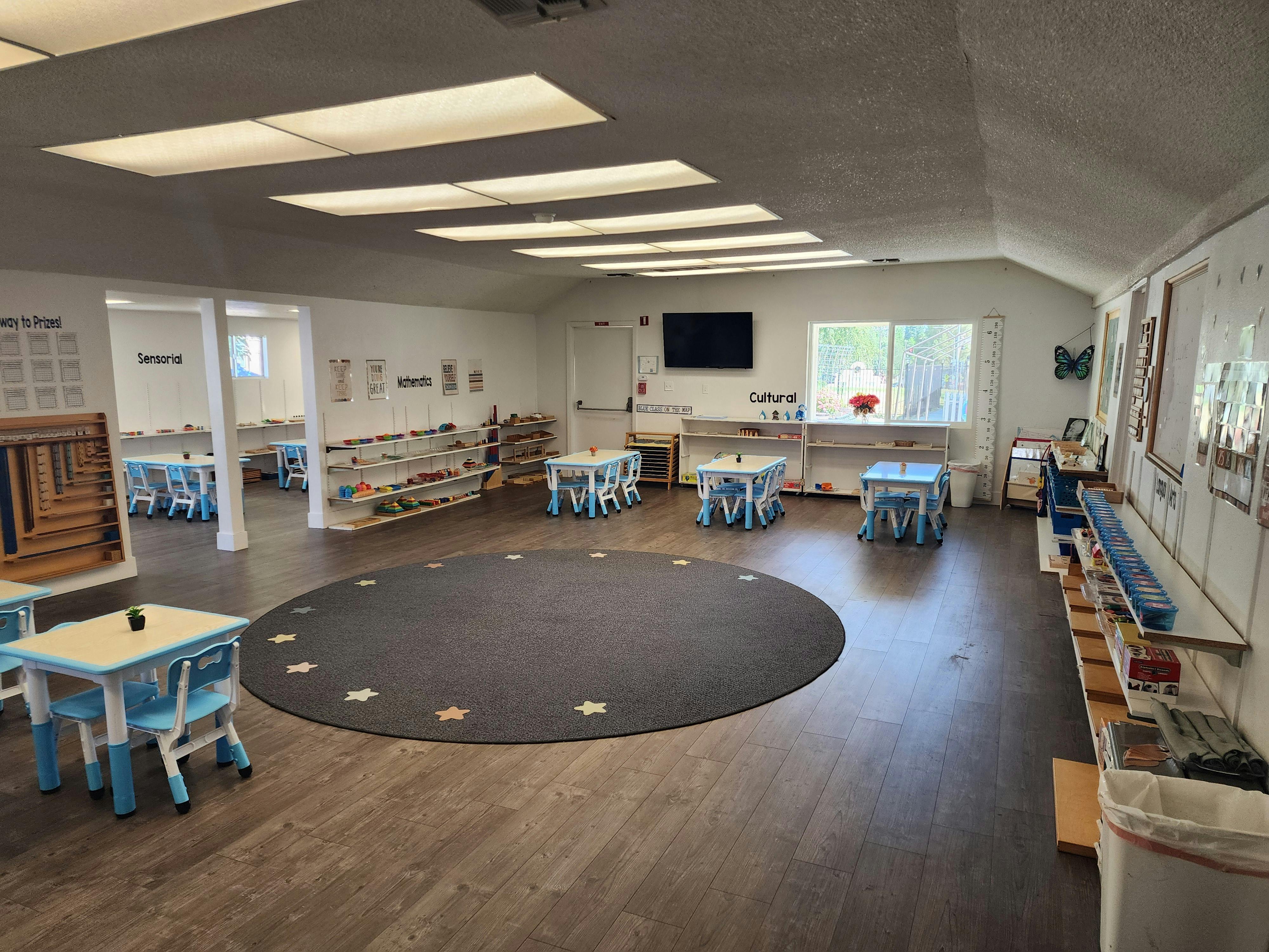 Classroom with small tables and chairs, a circular rug, and shelves
