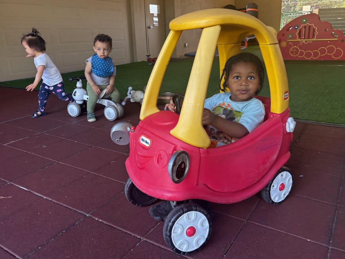 Child in a red toy car with two children in the background